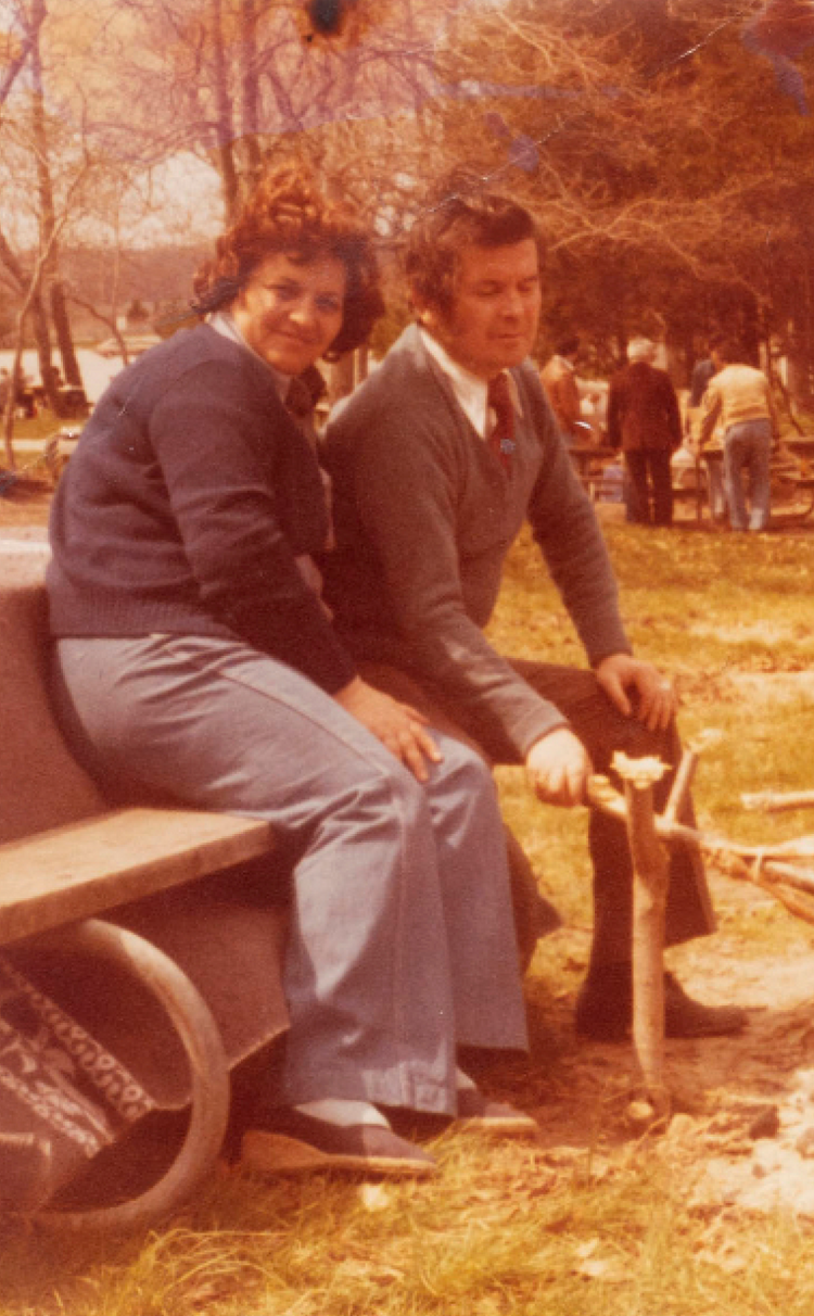 Couple posing for photo sitting on a park bench tending to a barbecue pit 