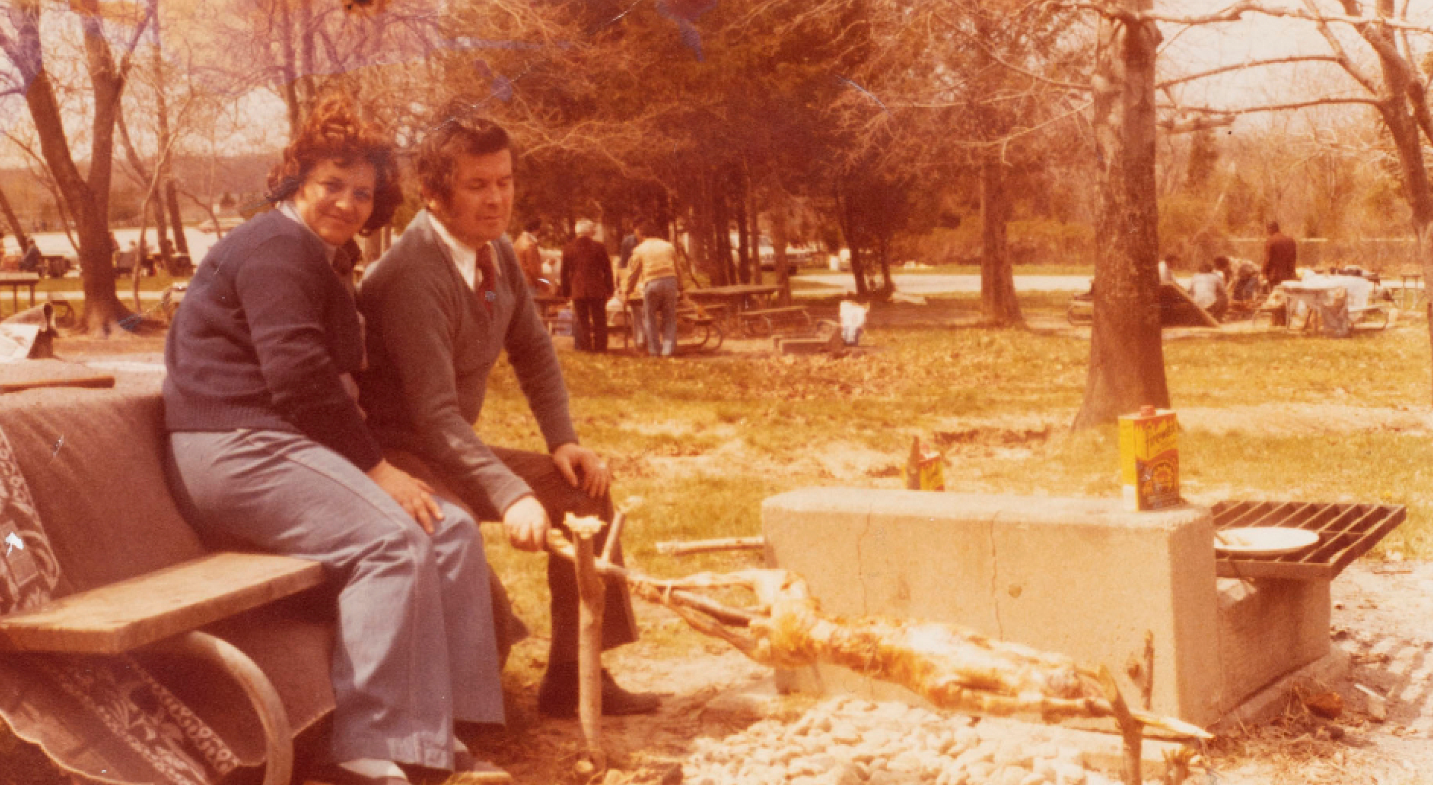 Couple posing for photo sitting on a park bench tending to a barbecue pit 