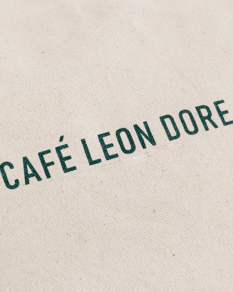 The Café Leon Dore Tote Bag features green text on a textured, off-white fabric background.