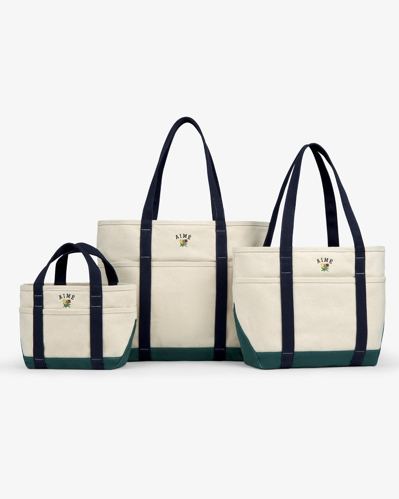 Large Crest Contrast Tote Bag: Canvas tote with green base, navy handles, and embroidered AIME logo on white.