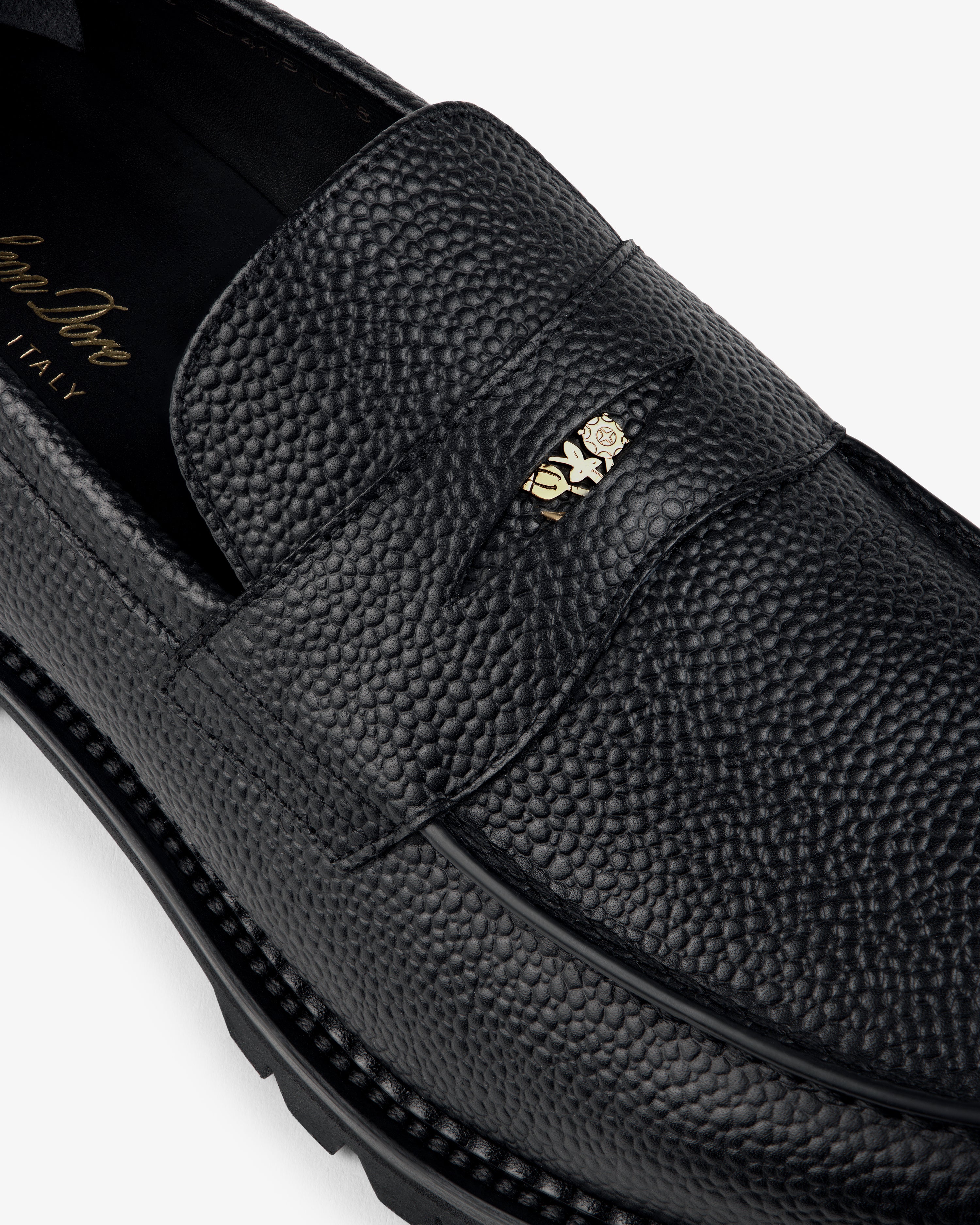 Close-up of the Lug Sole Queens Crest Loafer in black textured leather with a gold-tone metal strap detail.