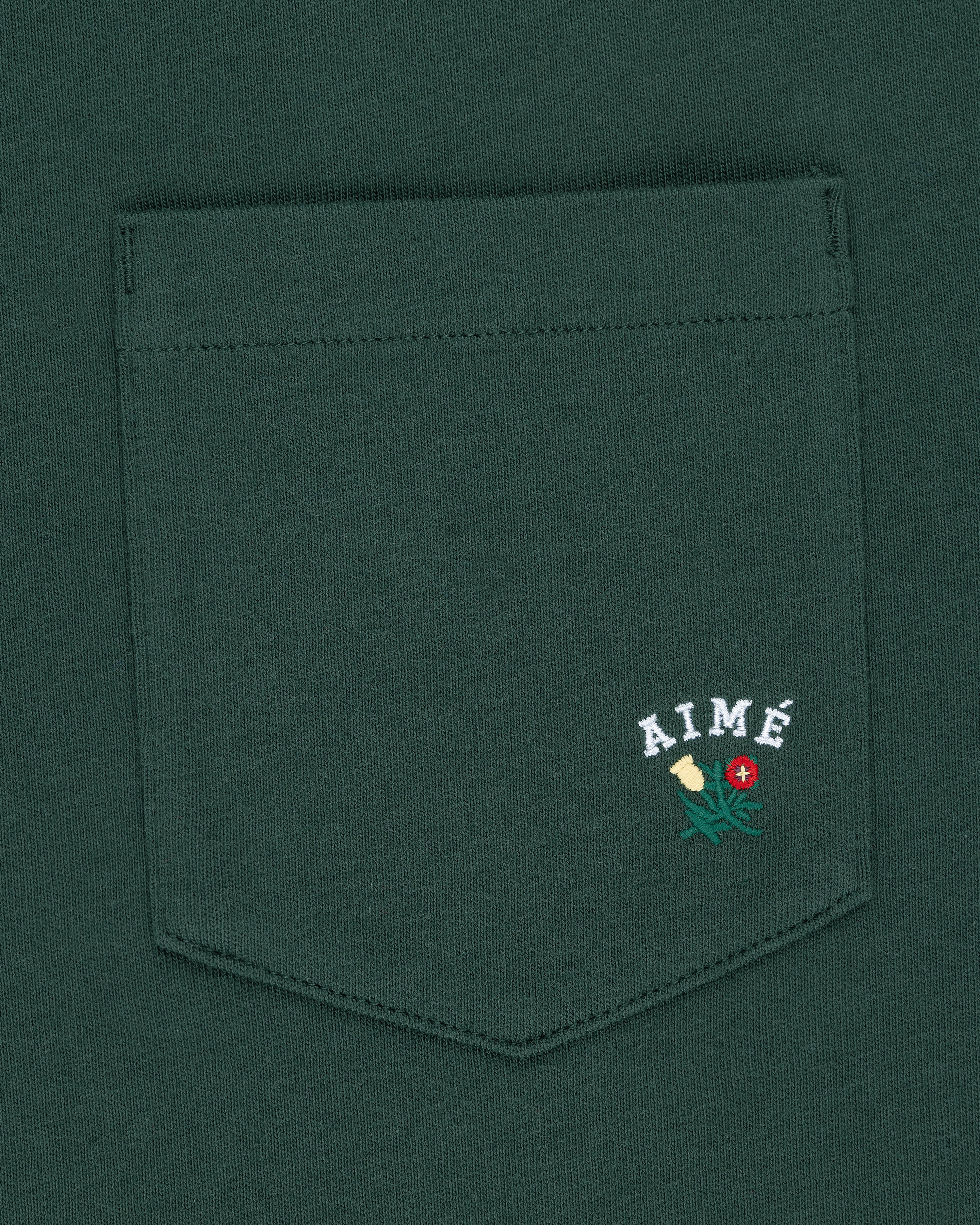 Crest Pocket Tee with AIMÉ text and an embroidered yellow flower, green leaves, and a red dot on the pocket.
