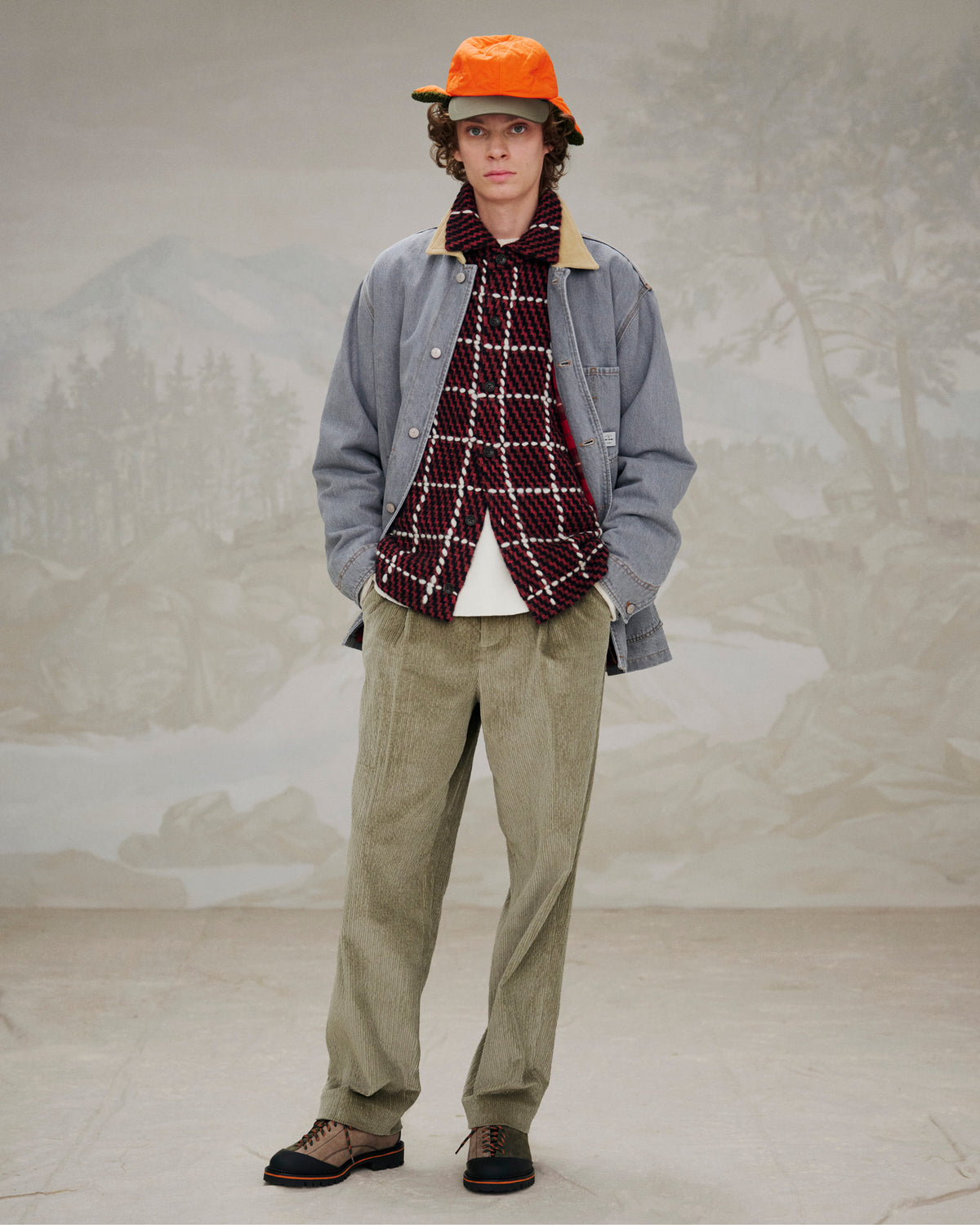 Person in orange cap, plaid jacket, grey shirt, tan corduroy pants, and Suede Derby Hiker stands by a painted backdrop.