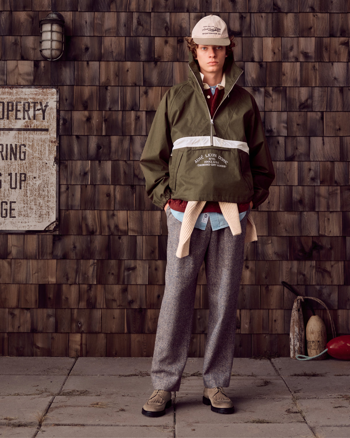 Person wearing Suede Lug Sole Derby, green jacket, gray trousers, and cap stands by a shingle wall with vintage props.