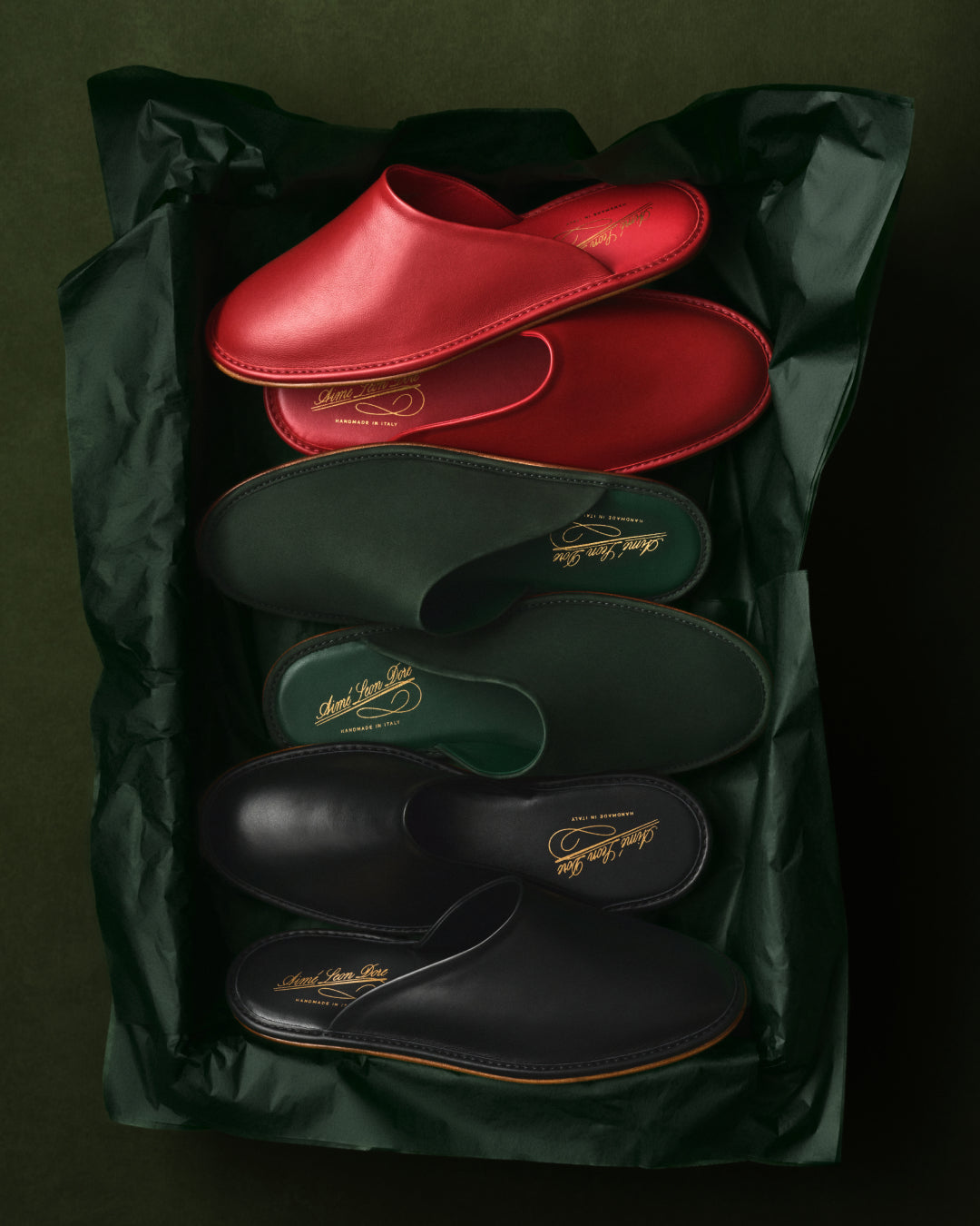 Three pairs of Leather Travel Slippers in red, green, and black displayed on dark green tissue paper.