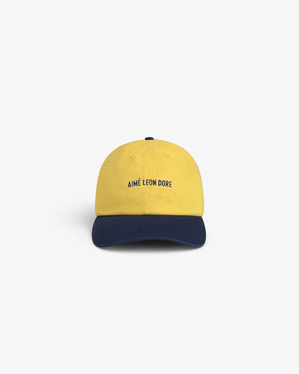 Pigment Dyed Logo Hat: yellow cap with AIMÉ LEON DORE front text and dark blue curved brim, on white background.