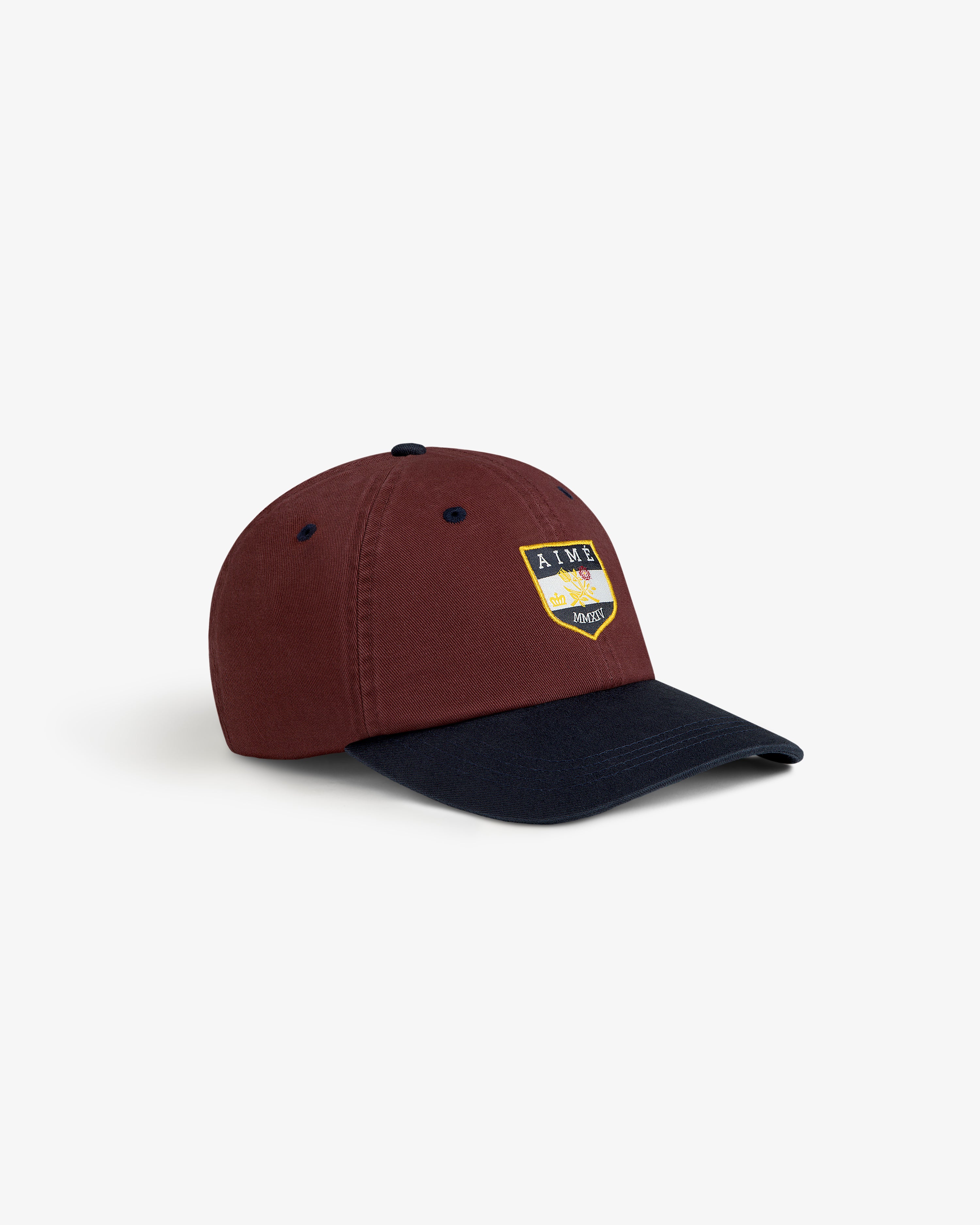 The Crest Shield Hat is maroon with a black brim and features a yellow, blue, and purple front patch.