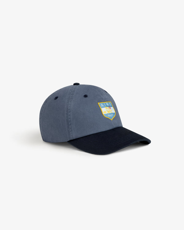 Crest Shield Hat: Blue cap with a curved black brim and yellow-blue crest patch on the front.