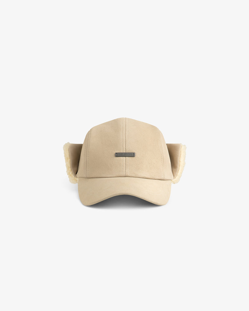 Beige suede Shearling Earflap Hat with fleece-lined earflaps and a rectangular metal logo plate on the front.