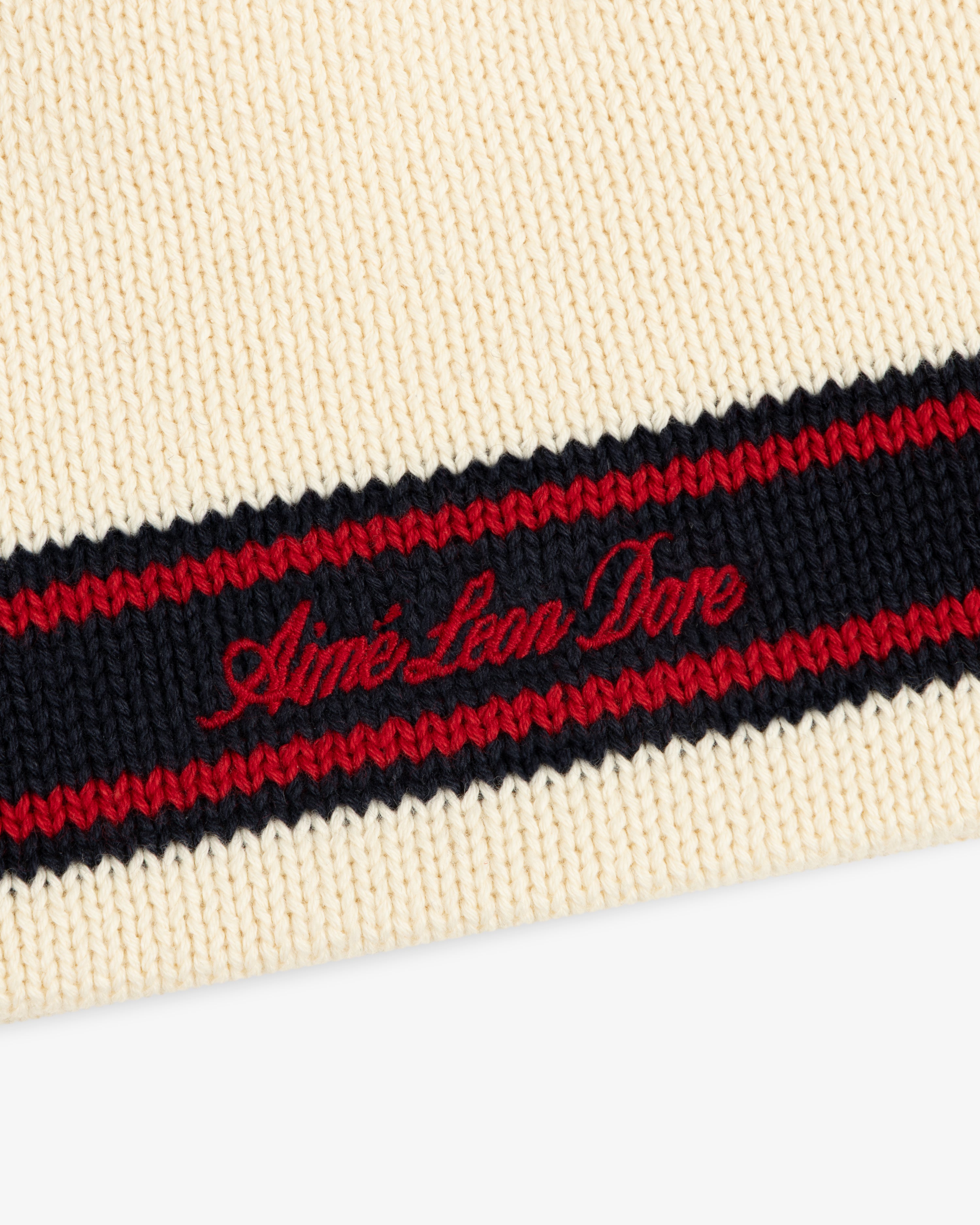 Close-up of the Stripe Knit Beanie in cream with black and red stripes, featuring red Aimé Leon Dore embroidery.