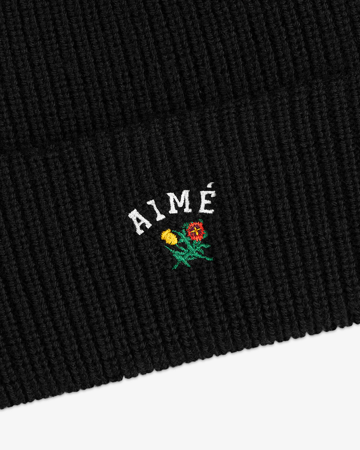 Crest Merino Wool Beanie in black ribbed knit with AIMÉ text and small yellow/red embroidered flowers underneath.