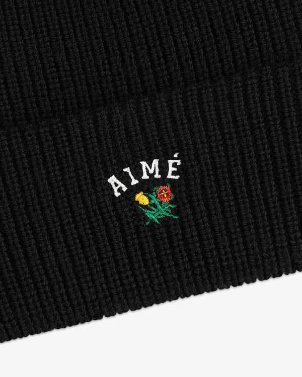 Crest Merino Wool Beanie in black ribbed knit with AIMÉ text and small yellow/red embroidered flowers underneath.