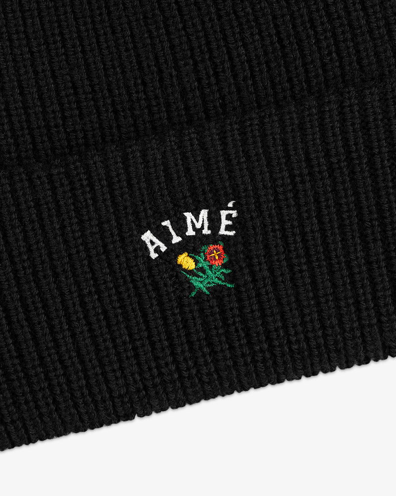 Crest Merino Wool Beanie in black ribbed knit with AIMÉ text and small yellow/red embroidered flowers underneath.
