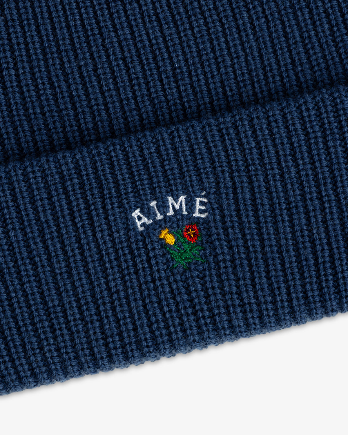 Close-up of the Crest Merino Wool Beanie in blue with AIME and an embroidered yellow and red flower on the cuff.