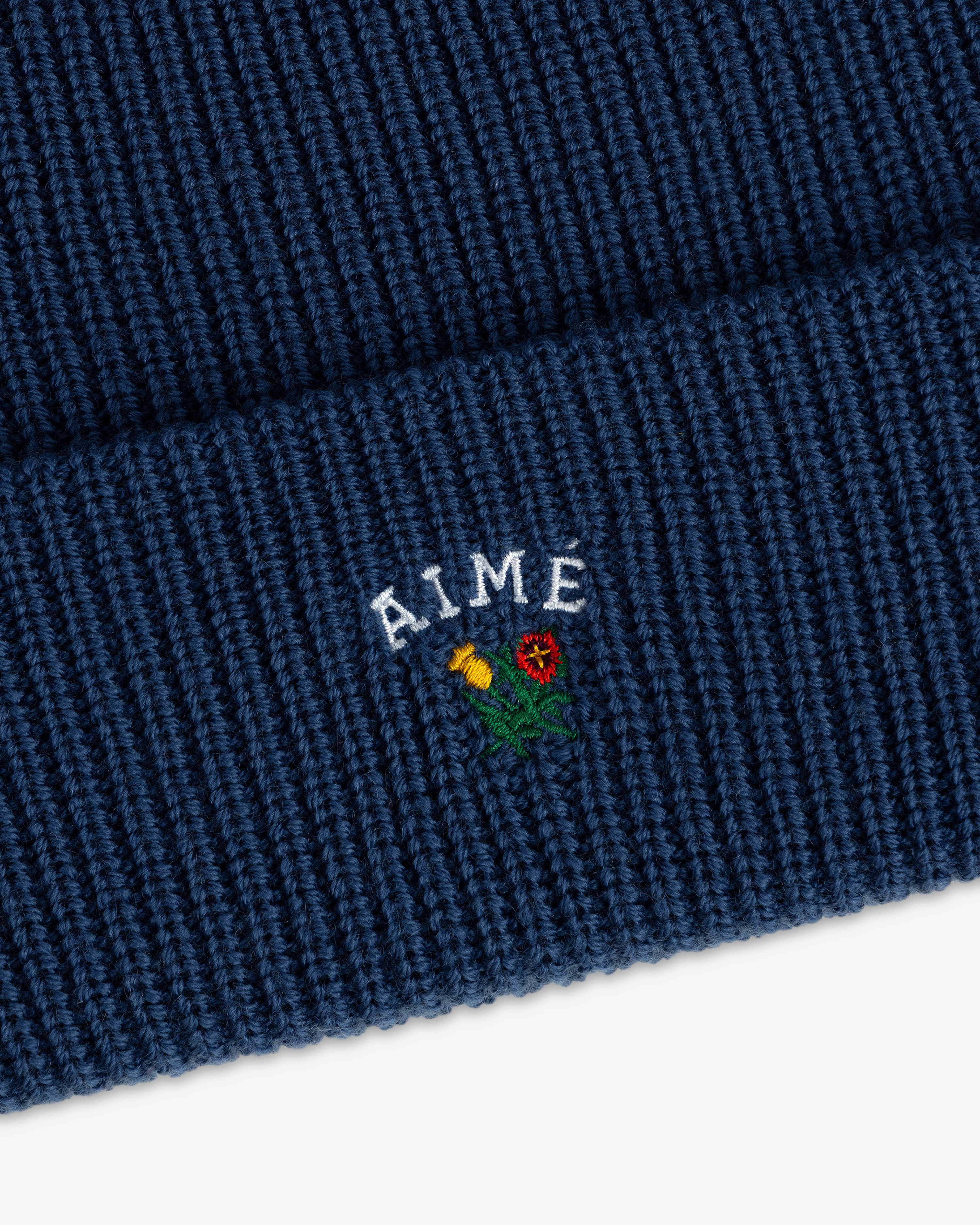 Close-up of the Crest Merino Wool Beanie in blue with AIME and an embroidered yellow and red flower on the cuff.