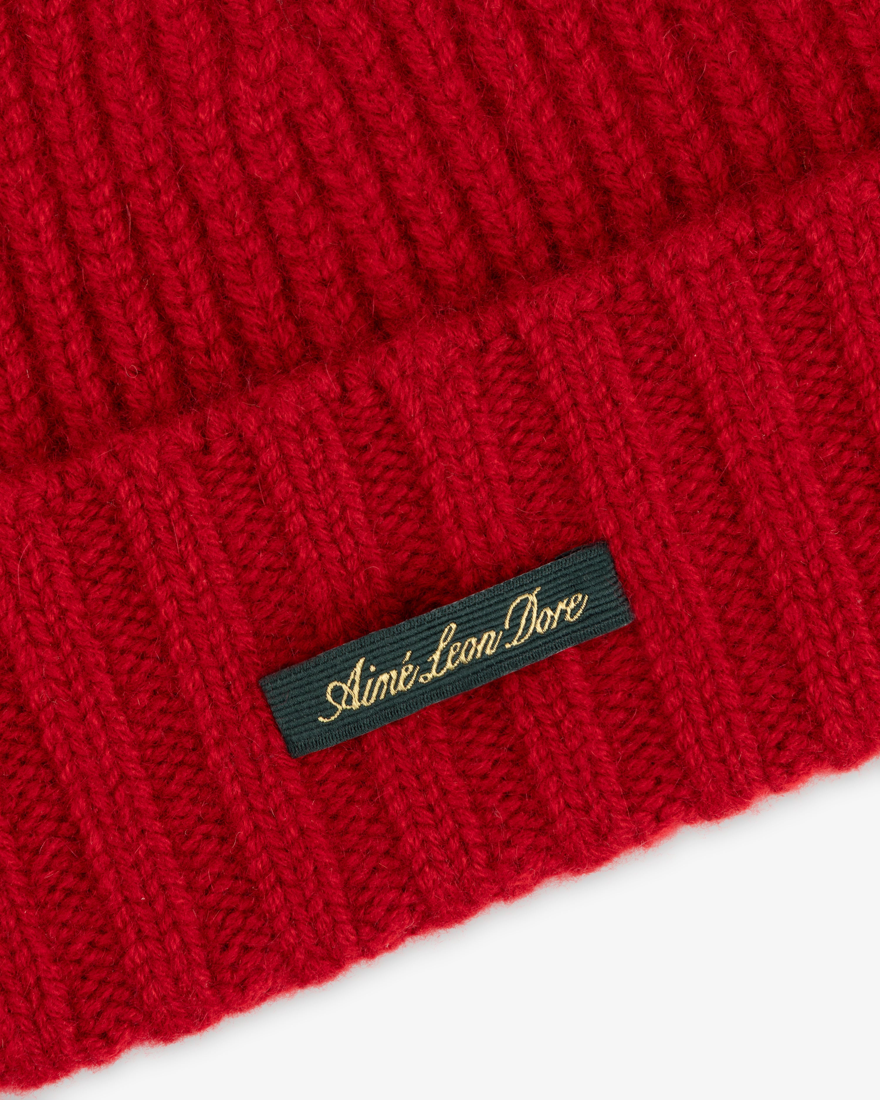 Close-up of a red ribbed Cashmere Beanie featuring a dark green tag embroidered with Aimé Leon Dore in gold script.