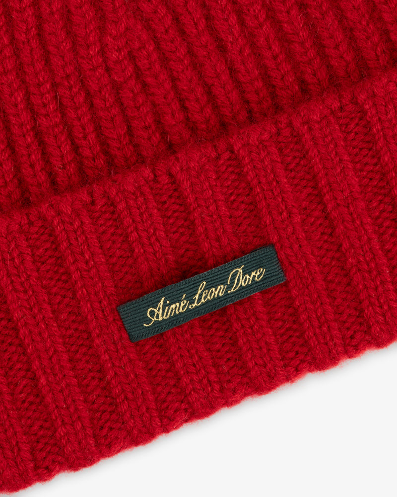Close-up of a red ribbed Cashmere Beanie featuring a dark green tag embroidered with Aimé Leon Dore in gold script.