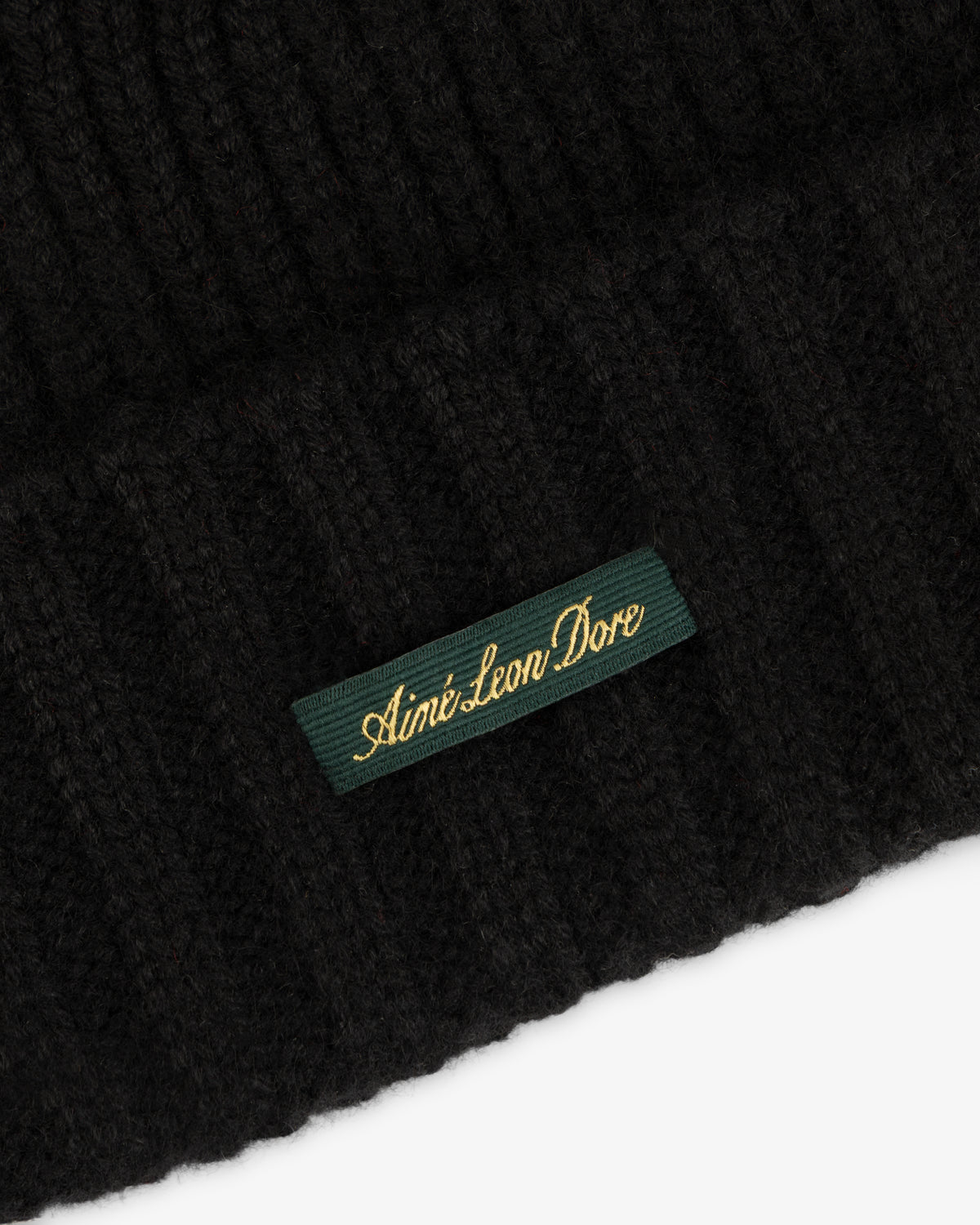 Close-up of a black ribbed knit Cashmere Beanie with a green woven label reading Aimé Leon Dore in gold script.