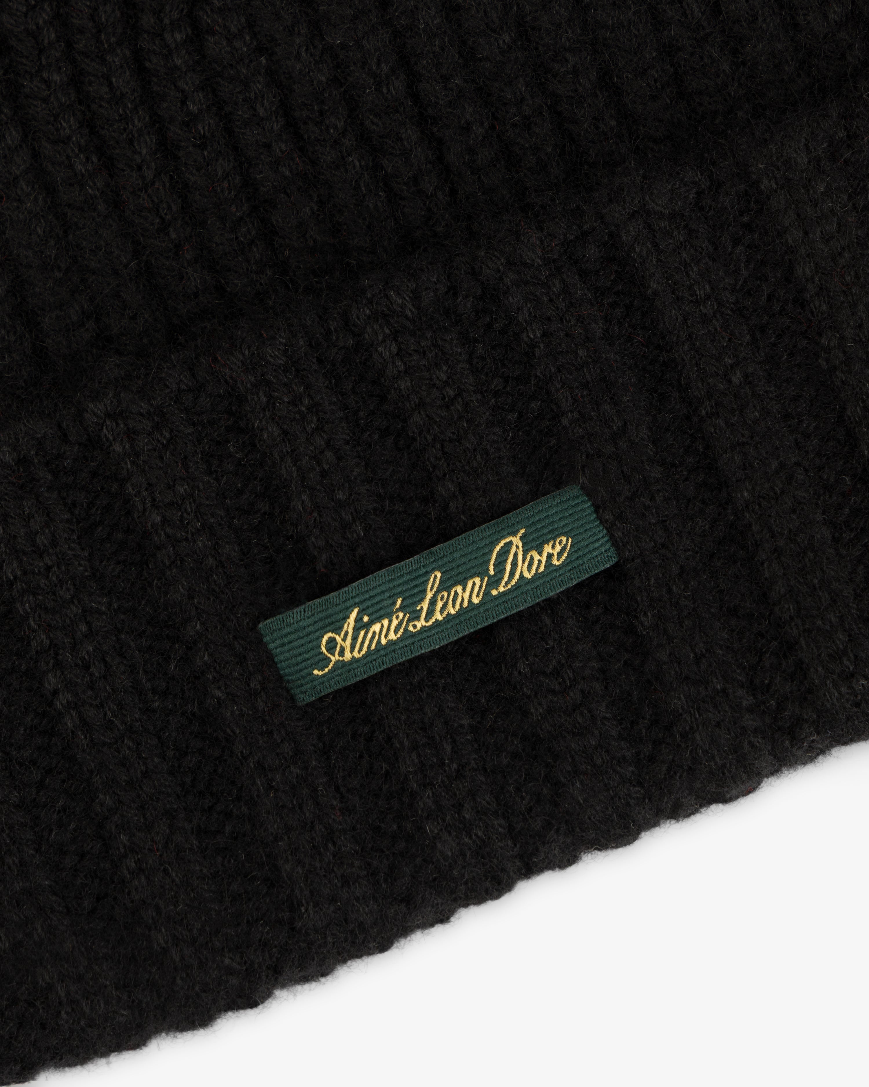 Close-up of a black ribbed knit Cashmere Beanie with a green woven label reading Aimé Leon Dore in gold script.