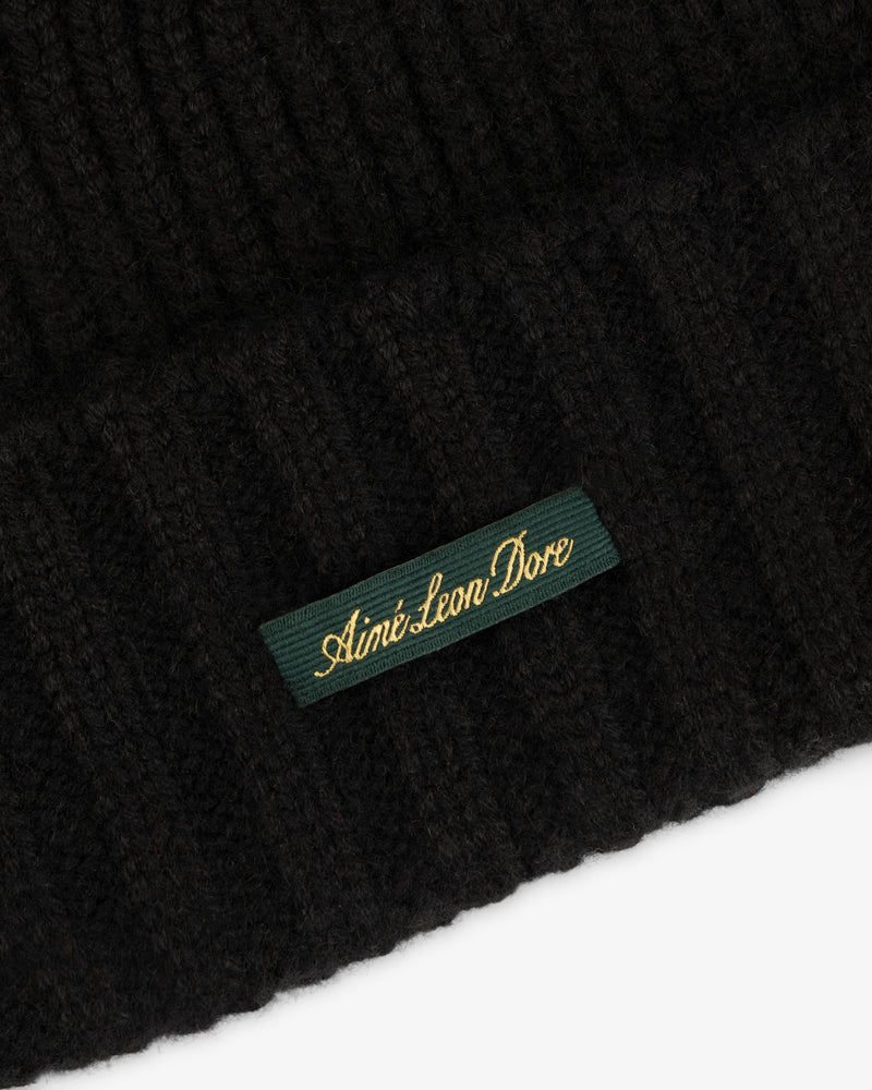 Close-up of a black ribbed knit Cashmere Beanie with a green woven label reading Aimé Leon Dore in gold script.