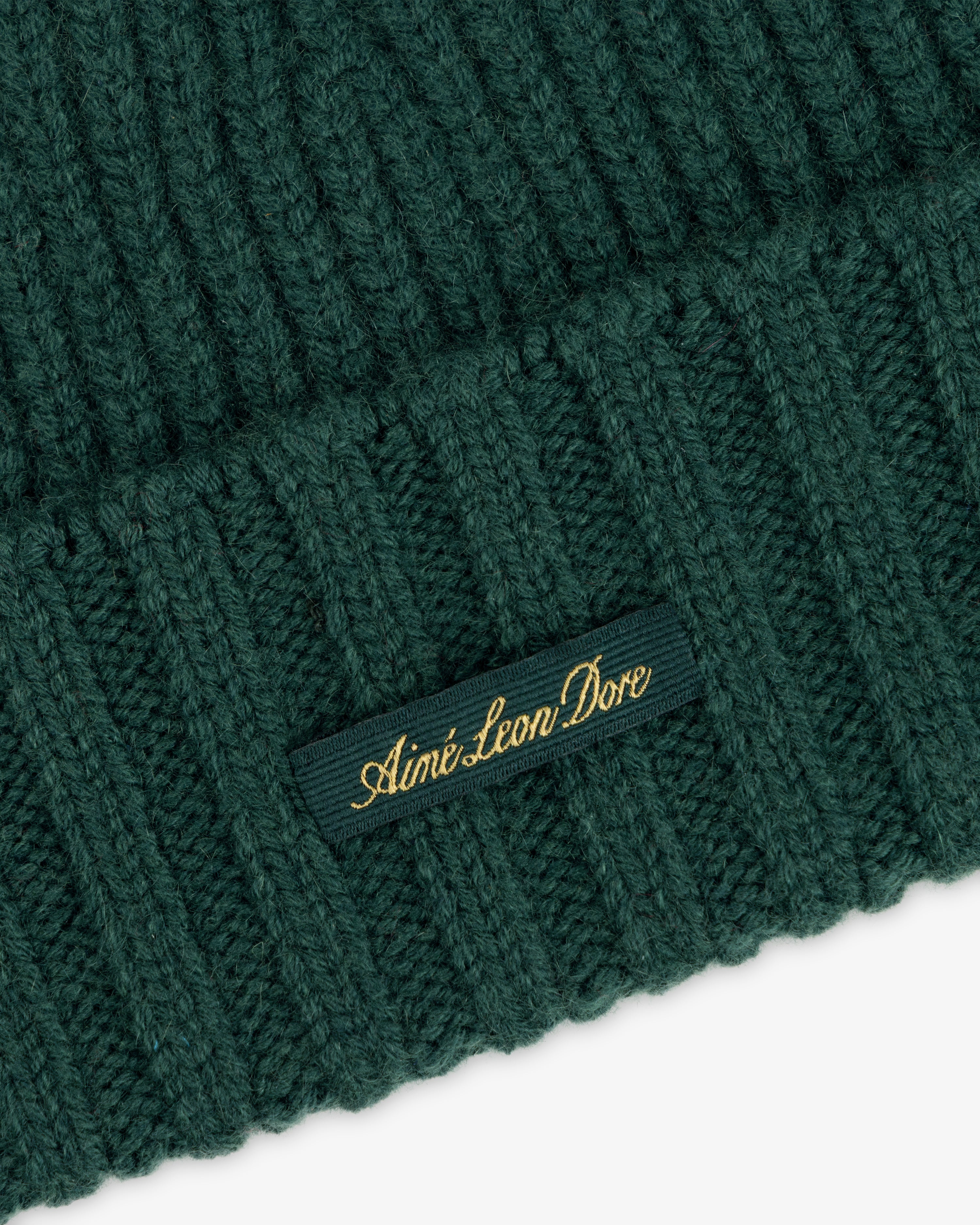 Dark green ribbed knit Cashmere Beanie with a small tag embroidered with Aimé Leon Dore in gold thread.