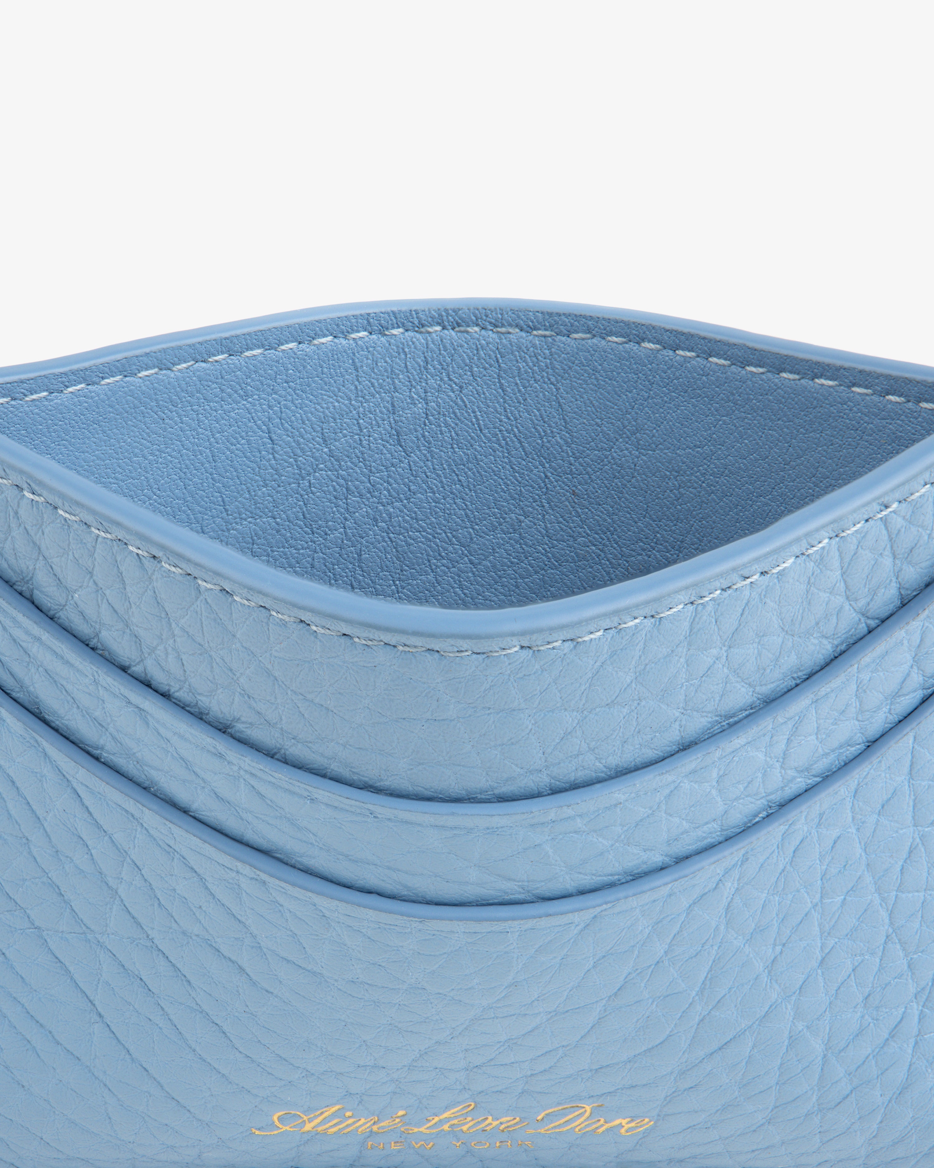 A close-up of the Tumbled Leather Card Holder in light blue leather with gold embossed text at the bottom.