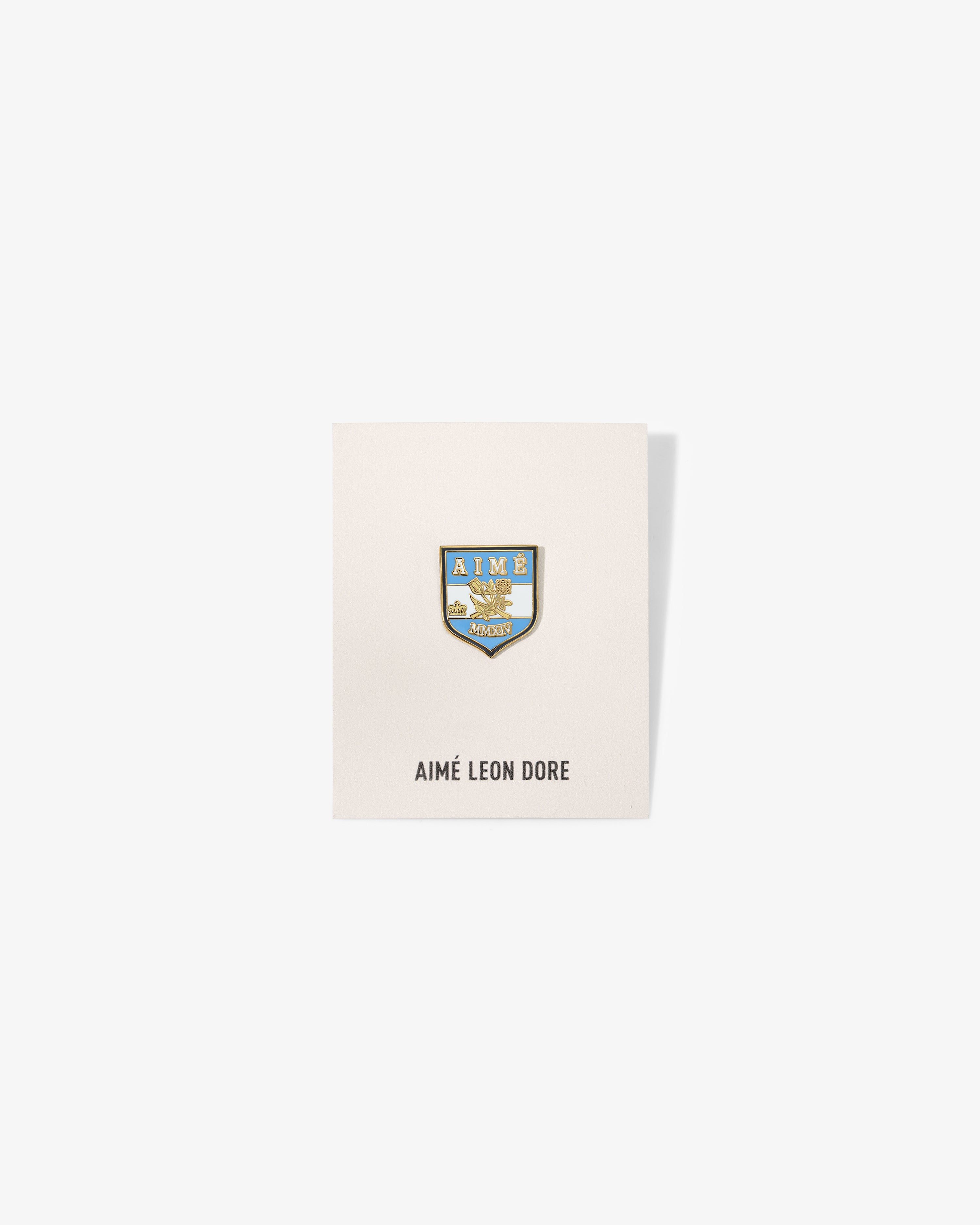 A blue and gold Graphic Pin on a white card labeled AIMÉ LEON DORE at the bottom.