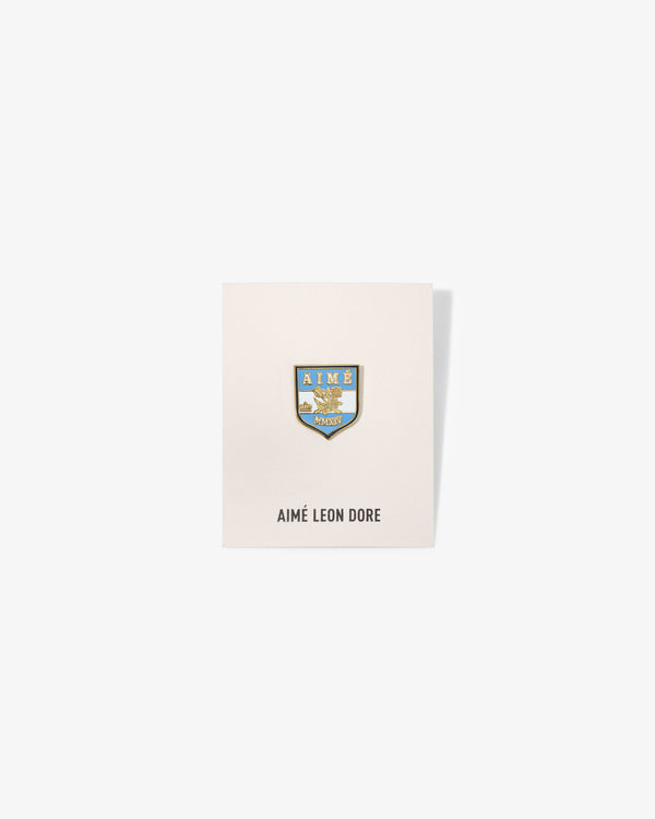 A blue and gold Graphic Pin on a white card labeled AIMÉ LEON DORE at the bottom.