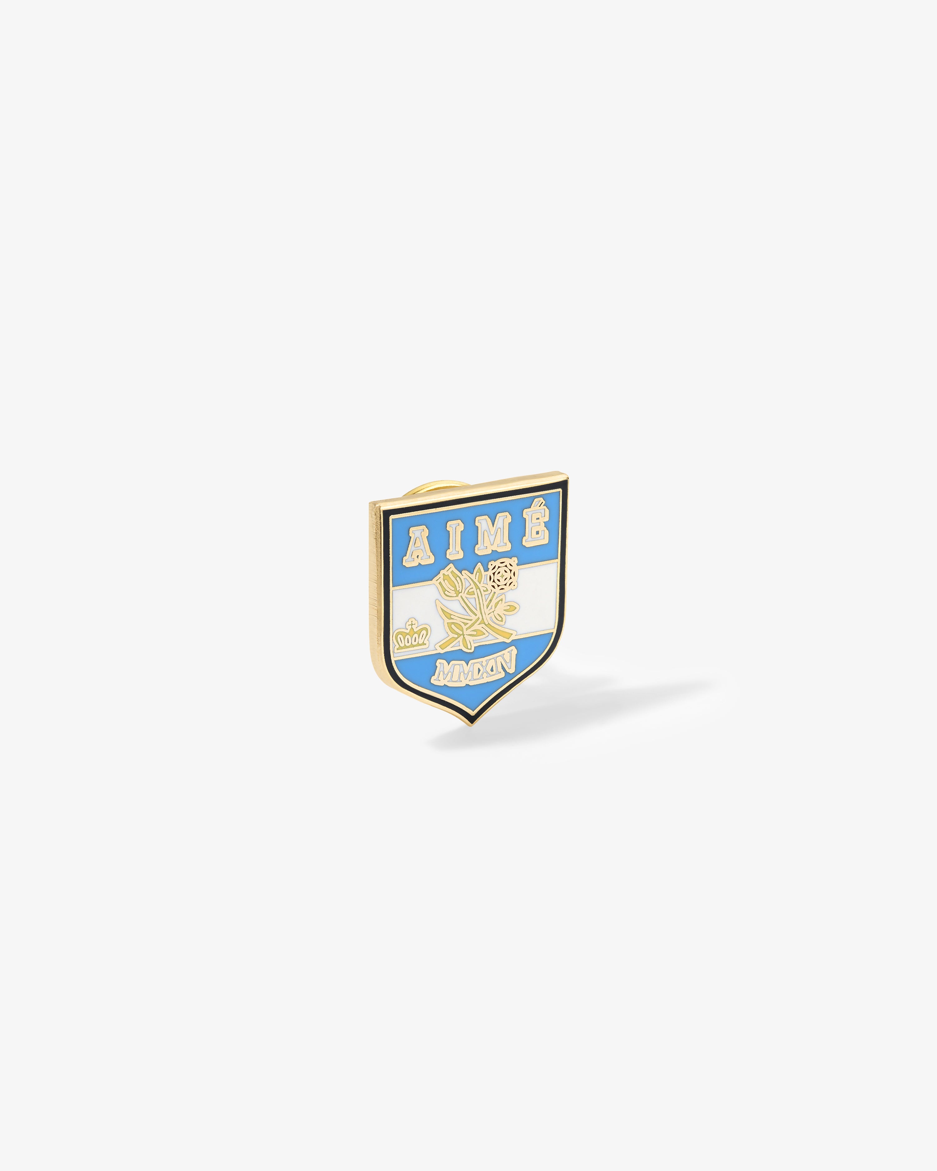 A gold and blue Graphic Pin with AIMÉ, floral design, and MONON lettering on a white shield background.