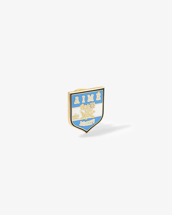 A gold and blue Graphic Pin with AIMÉ, floral design, and MONON lettering on a white shield background.