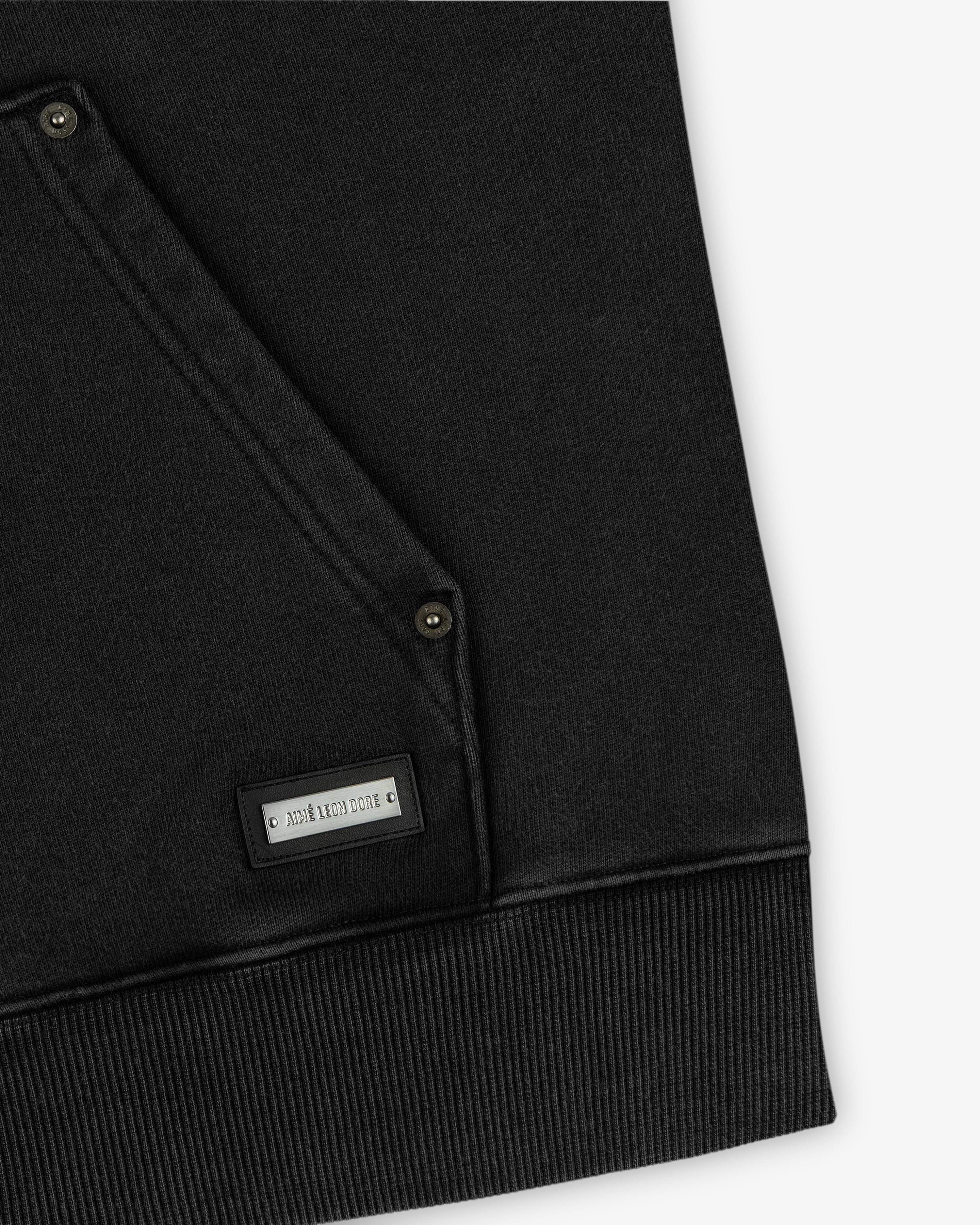 Close-up of a black Washed Full Zip Hoodie pocket with metal rivets and a branded rectangular metal plate.