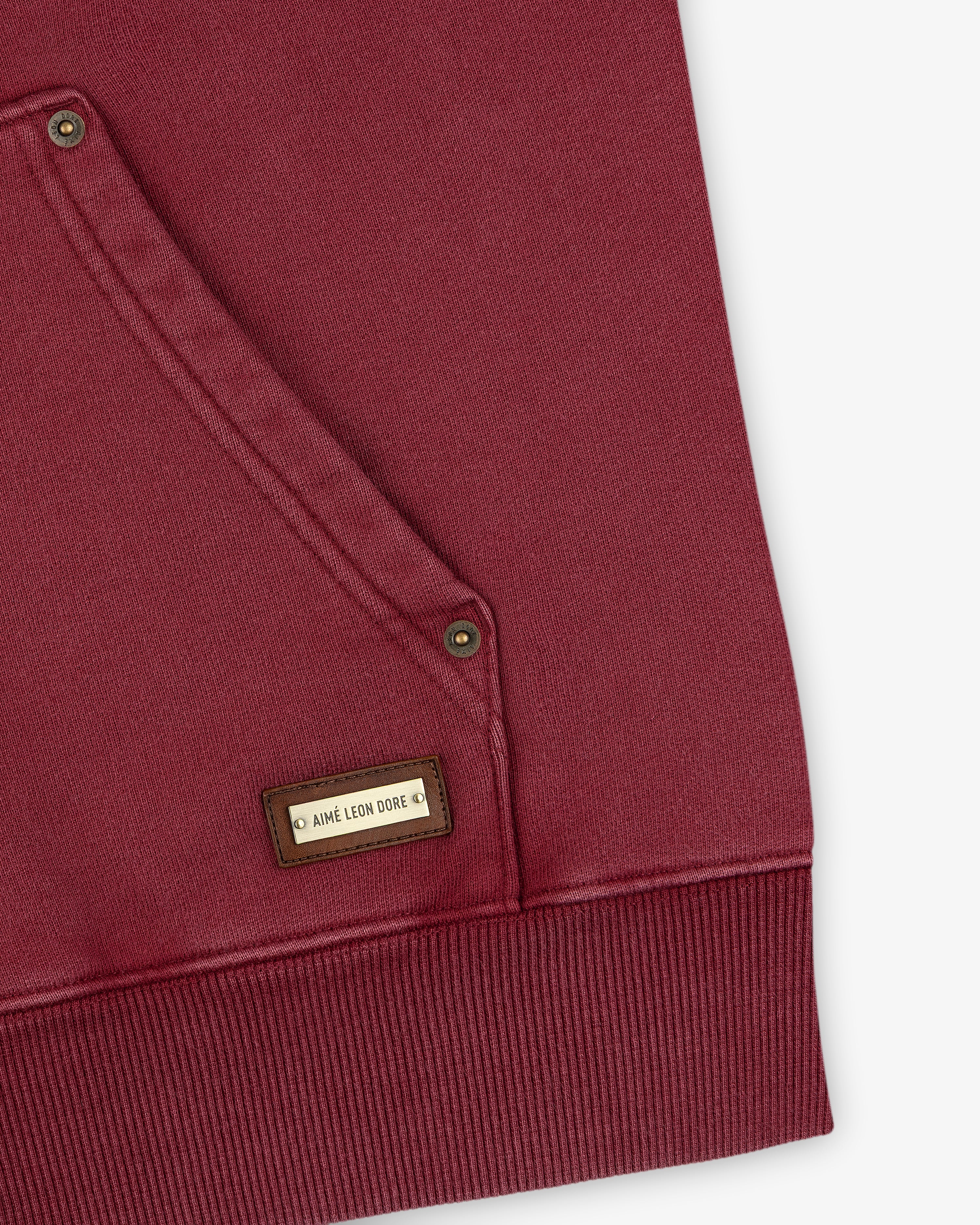 Close-up of a maroon Washed Full Zip Hoodie with a front pocket featuring metallic rivets and a rectangular brand tag.