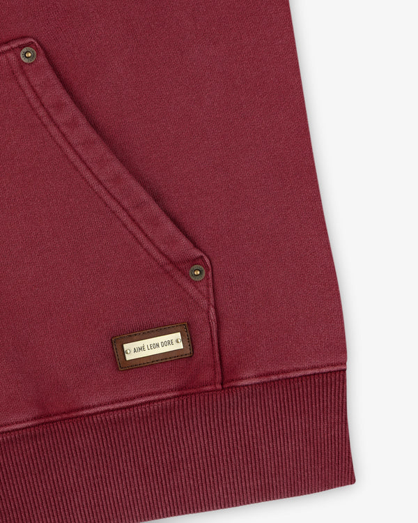 Close-up of a maroon Washed Full Zip Hoodie with a front pocket featuring metallic rivets and a rectangular brand tag.