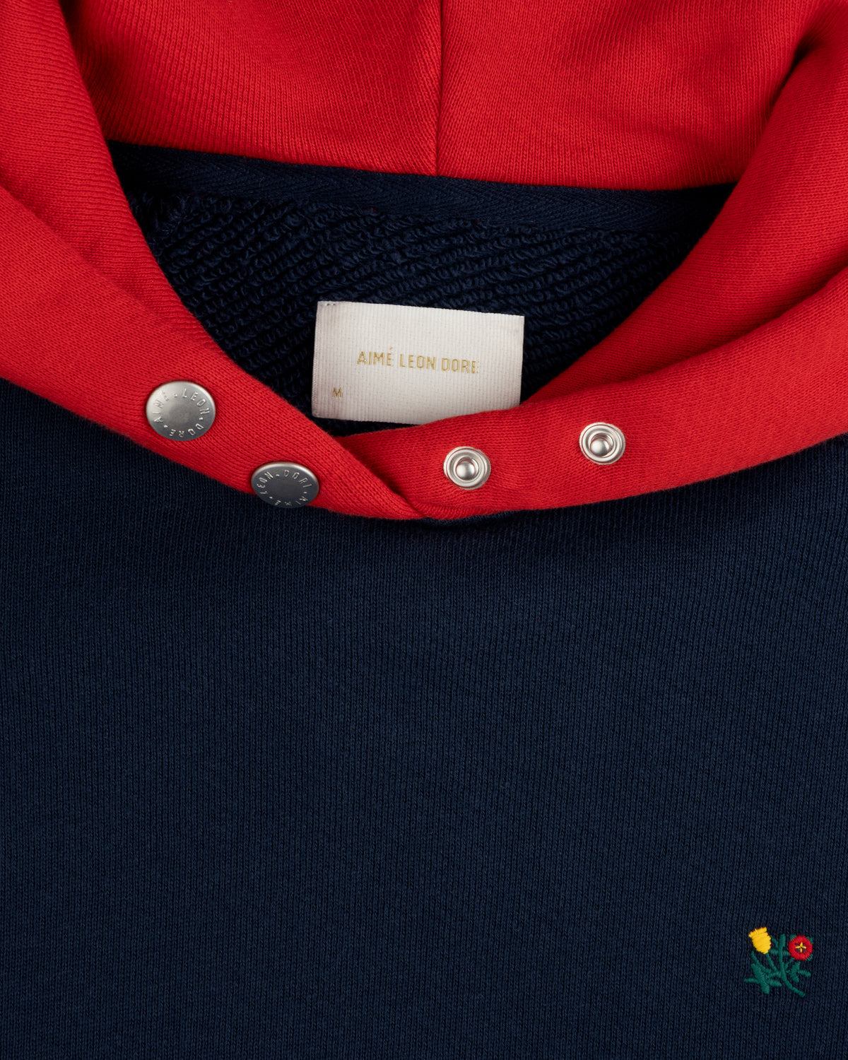 Colorblock Crest Snap Hoodie: navy hoodie with a red hood, snap-button collar, and small floral embroidery near the hem.
