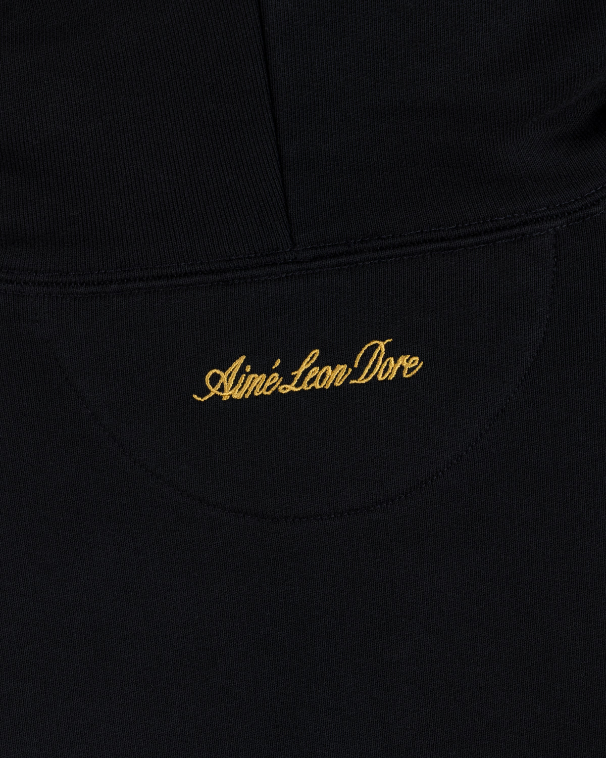 Black fabric of the ALD / Olympiacos F.C. Full Zip Hoodie with Aimé Leon Dore embroidered in gold script.