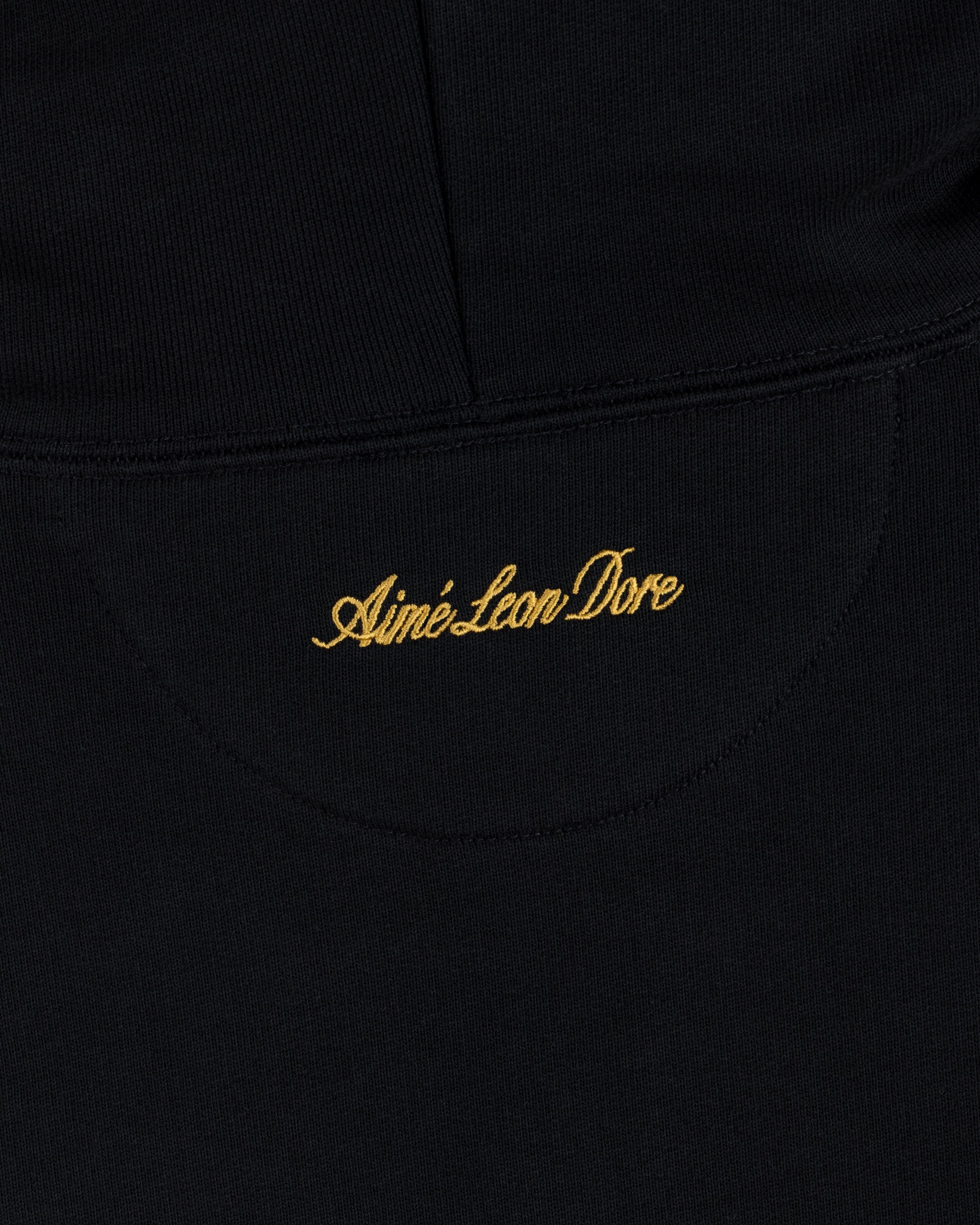 Black fabric of the ALD / Olympiacos F.C. Full Zip Hoodie with Aimé Leon Dore embroidered in gold script.