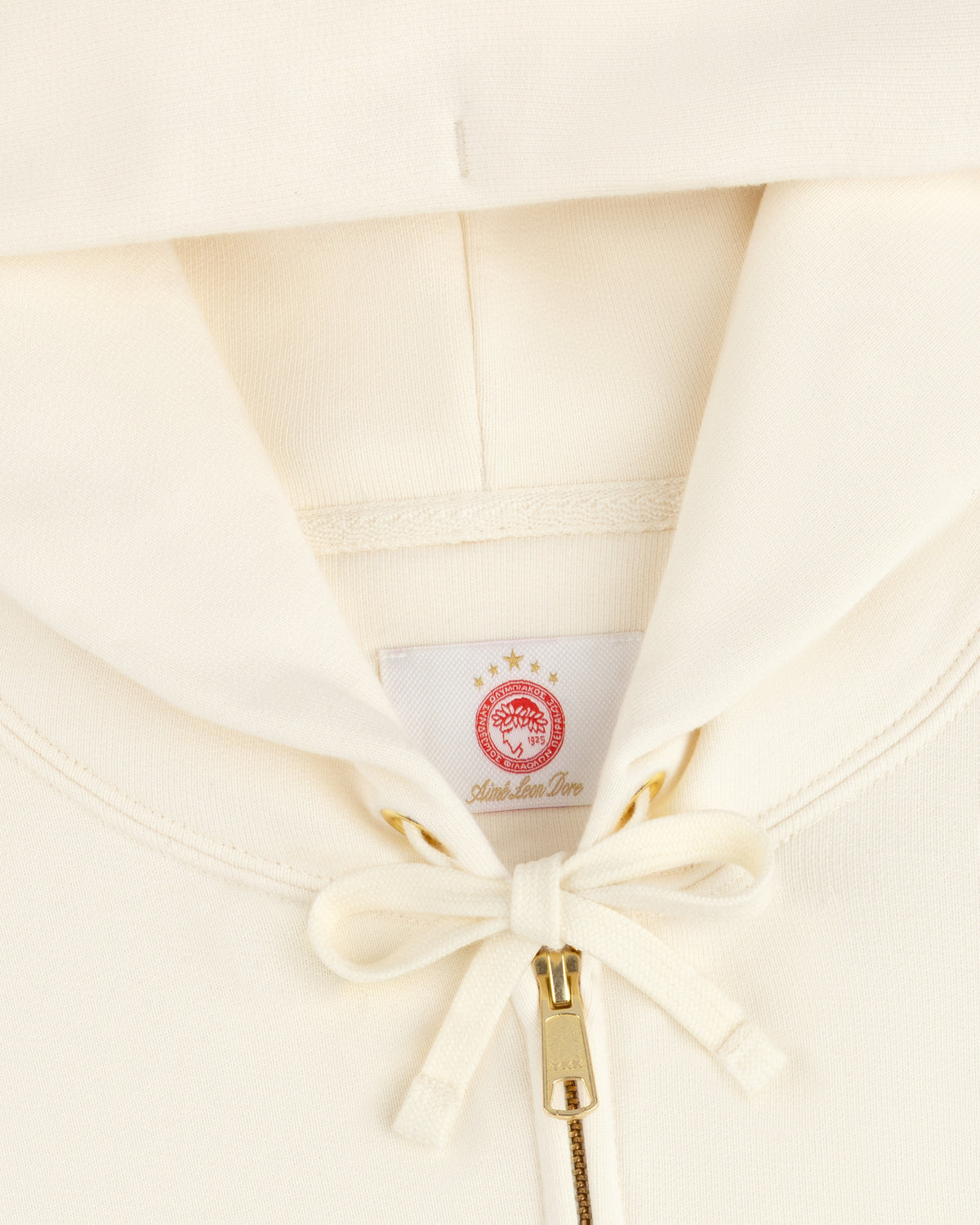 Close-up of a cream-colored ALD / Olympiacos F.C. Full Zip Hoodie with a red laurel wreath label and gold zipper.