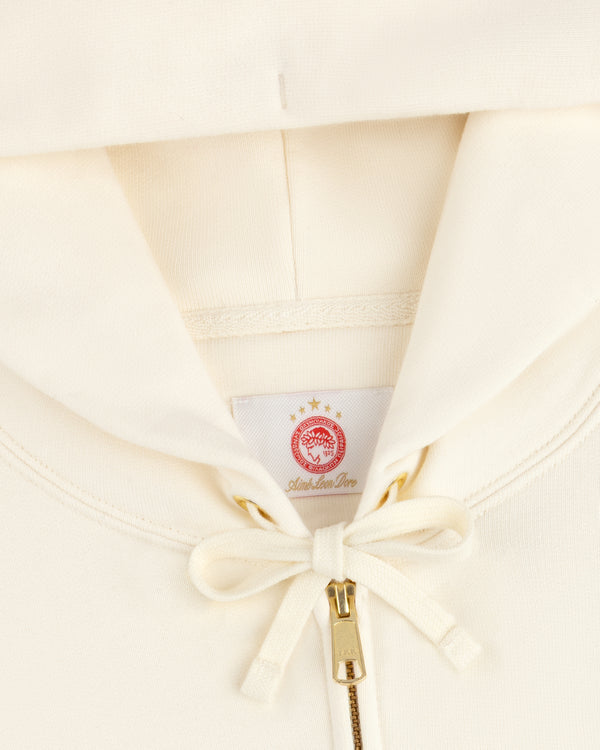 Close-up of a cream-colored ALD / Olympiacos F.C. Full Zip Hoodie with a red laurel wreath label and gold zipper.