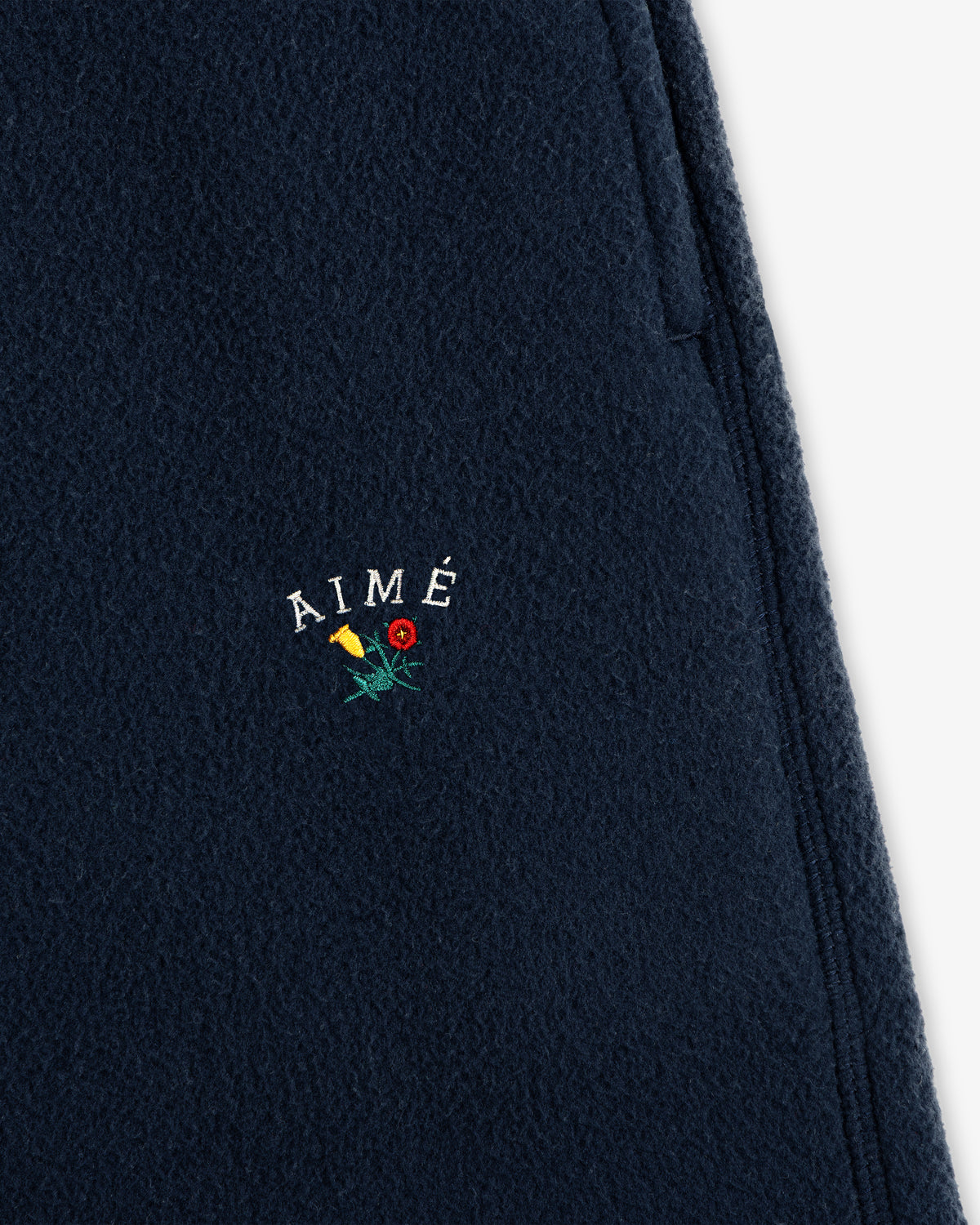 Crest Fleece Sweatpant in navy with AIMÉ and yellow/red flower embroidery above the zipper pocket.