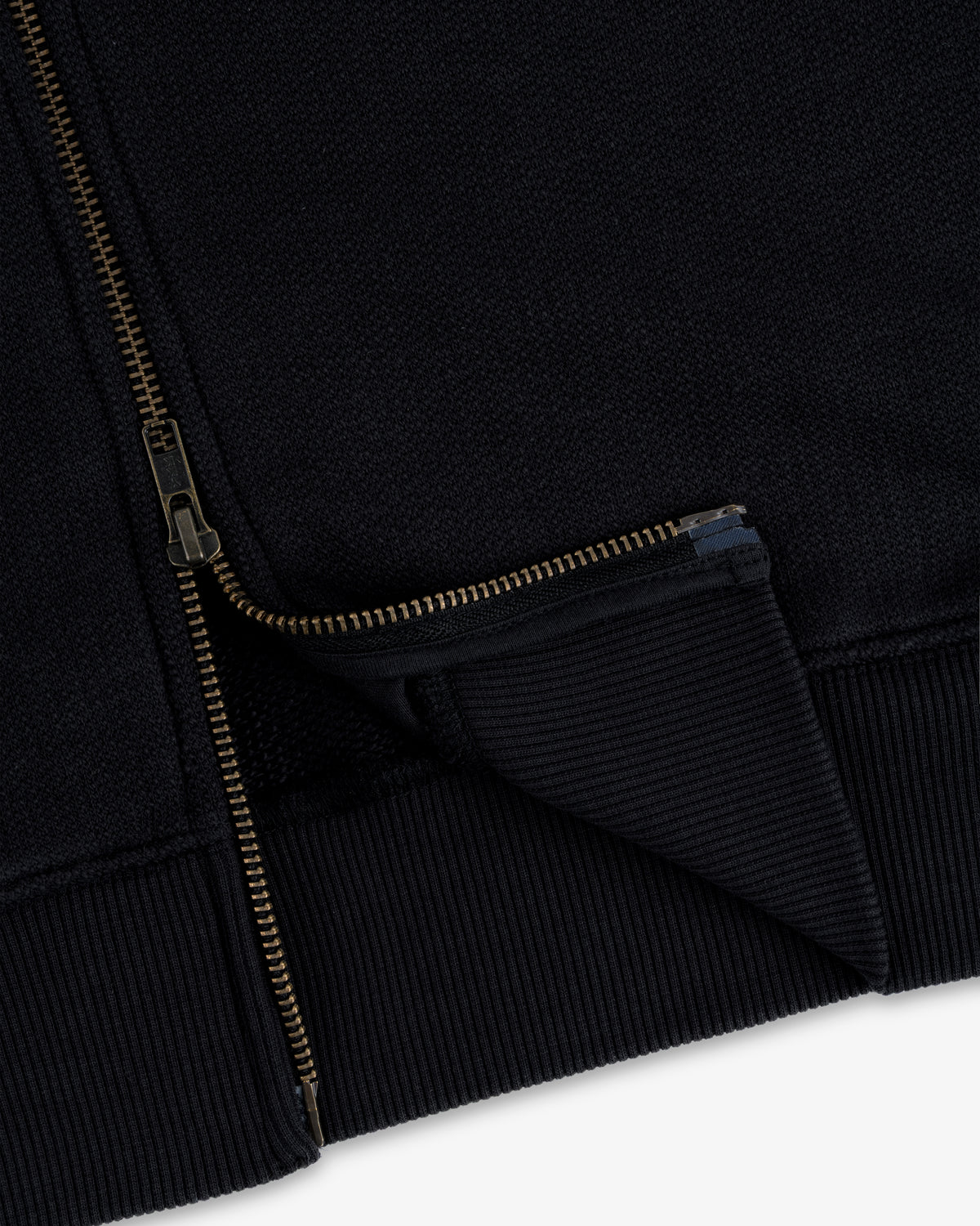Close-up of the Saint George Track Jacket featuring a gold zipper and ribbed hem on black fabric.