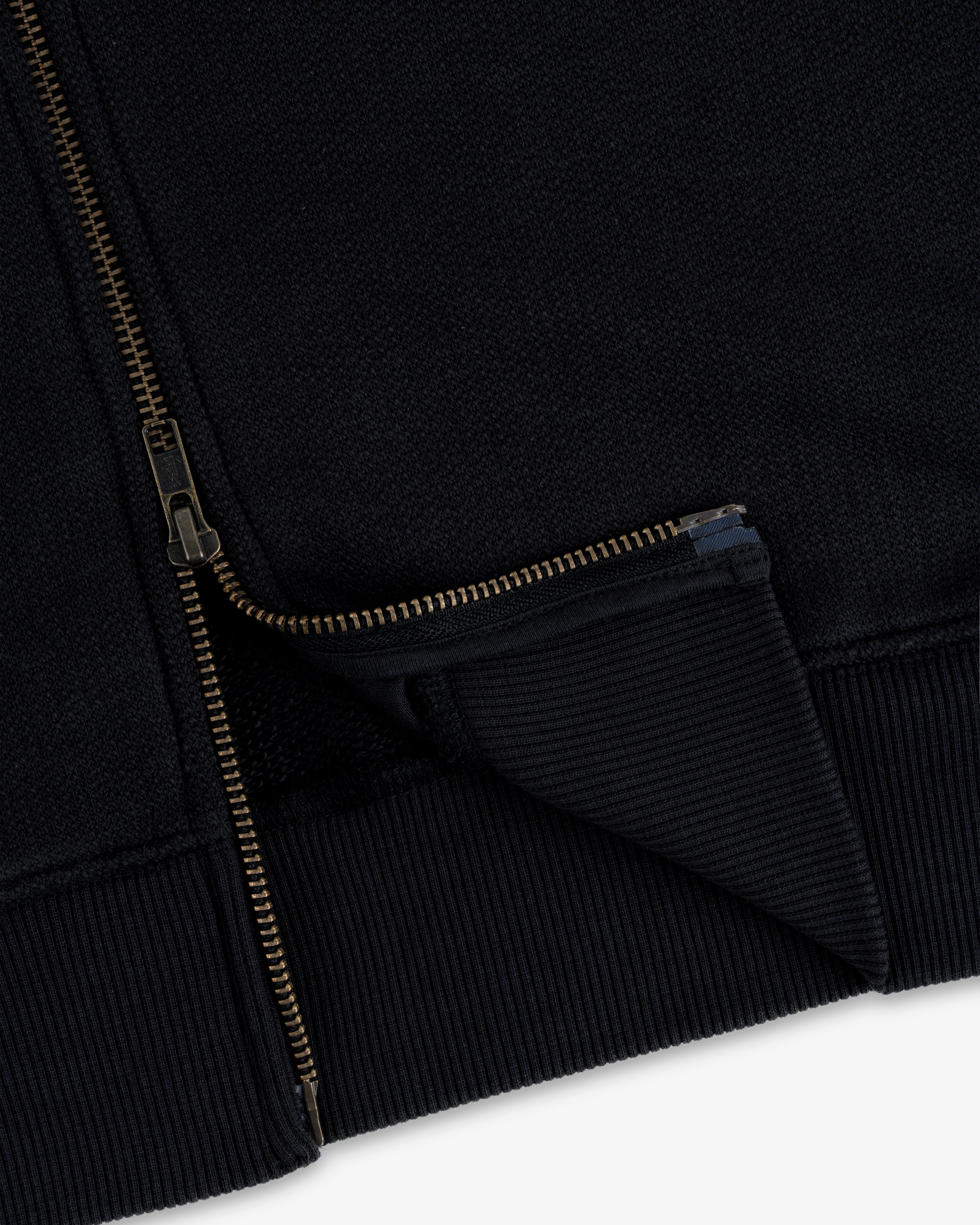 Close-up of the Saint George Track Jacket featuring a gold zipper and ribbed hem on black fabric.
