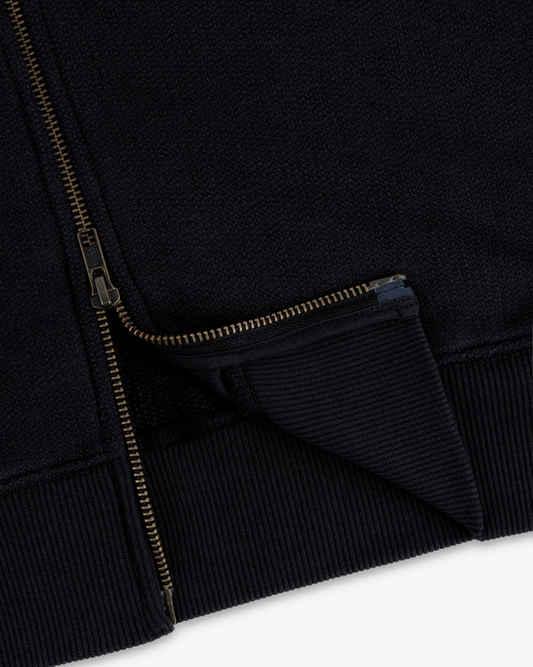 Close-up of the Saint George Track Jacket featuring a gold zipper and ribbed hem on black fabric.