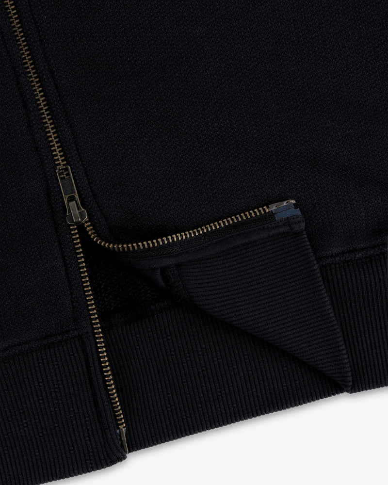 Close-up of the Saint George Track Jacket featuring a gold zipper and ribbed hem on black fabric.