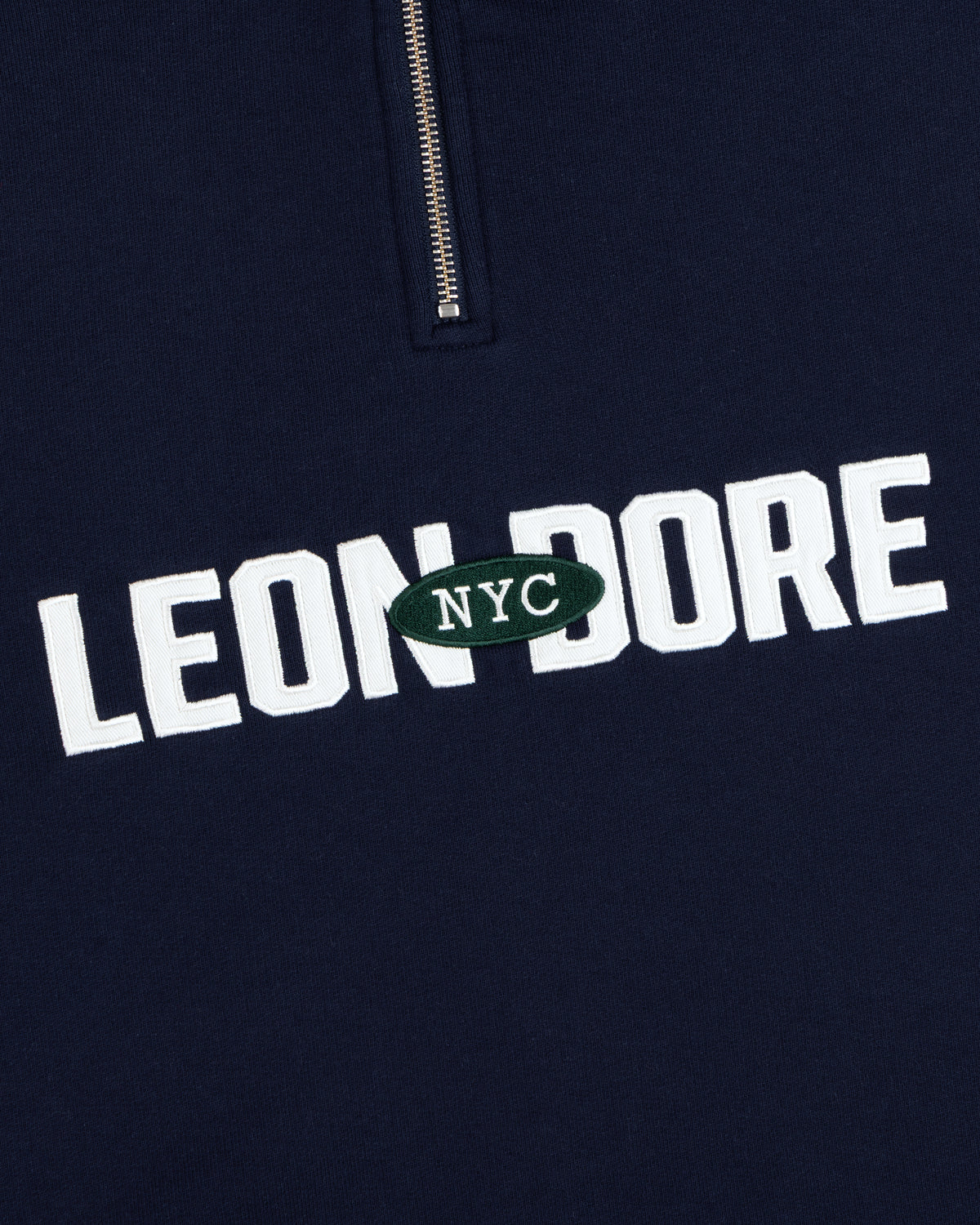 Aimé Souvenir Quarter Zip: Navy fabric, LEON DORE text, green oval NYC patch beneath a partial silver zipper.
