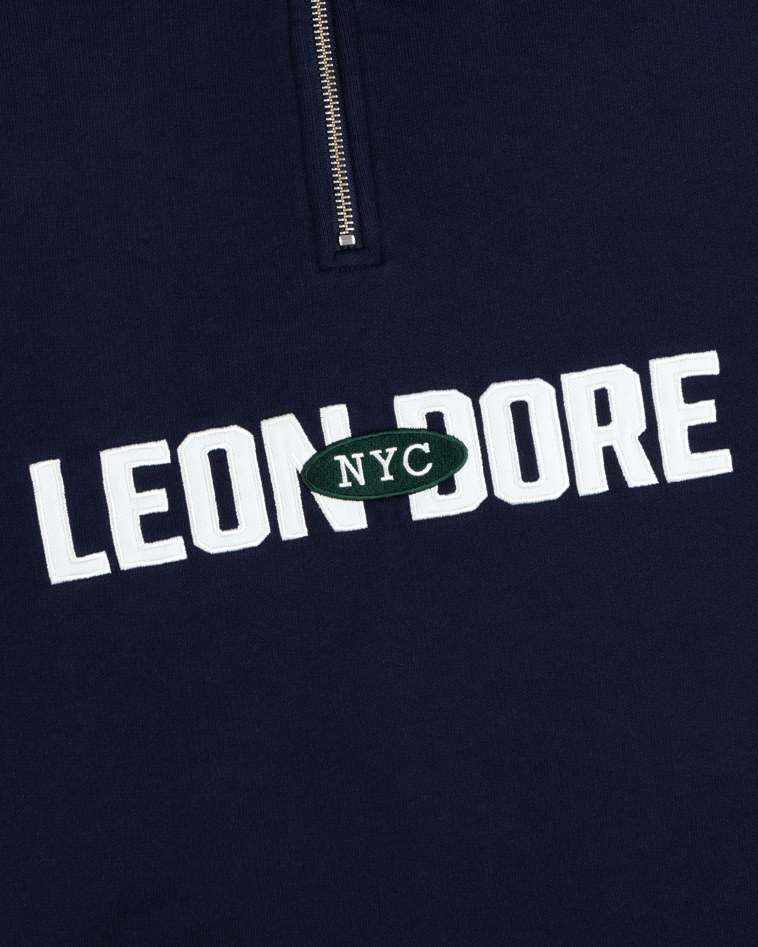 Aimé Souvenir Quarter Zip: Navy fabric, LEON DORE text, green oval NYC patch beneath a partial silver zipper.
