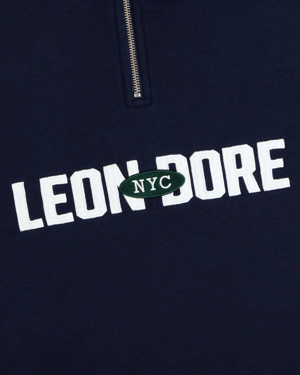 Aimé Souvenir Quarter Zip: Navy fabric, LEON DORE text, green oval NYC patch beneath a partial silver zipper.