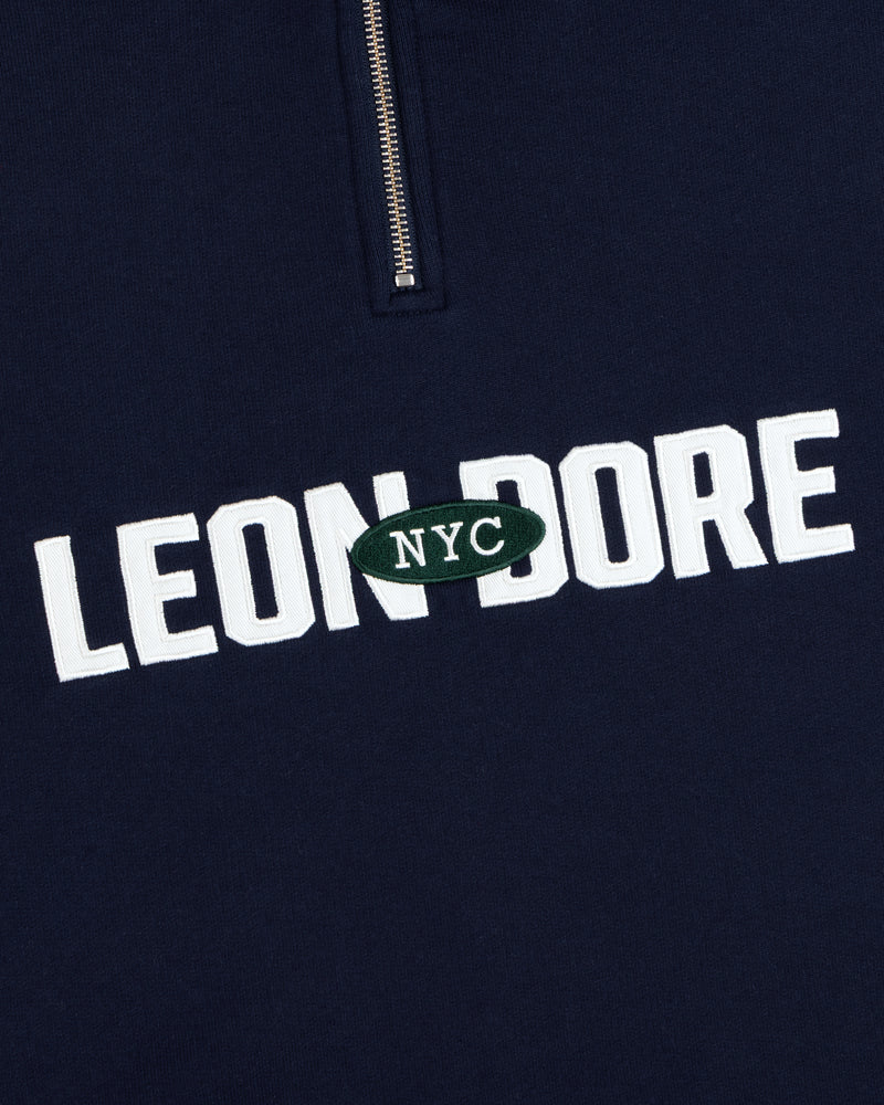 Aimé Souvenir Quarter Zip: Navy fabric, LEON DORE text, green oval NYC patch beneath a partial silver zipper.