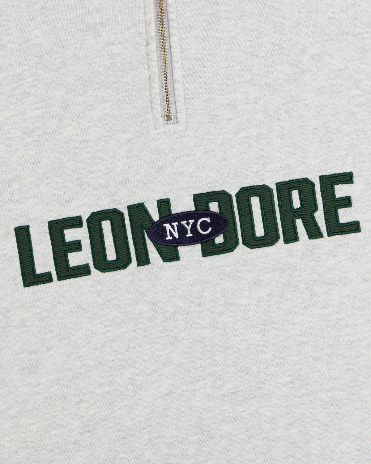 Close-up of Aimé Souvenir Quarter Zip’s light gray fabric, LEON DORE in green, NYC in white on a navy oval.