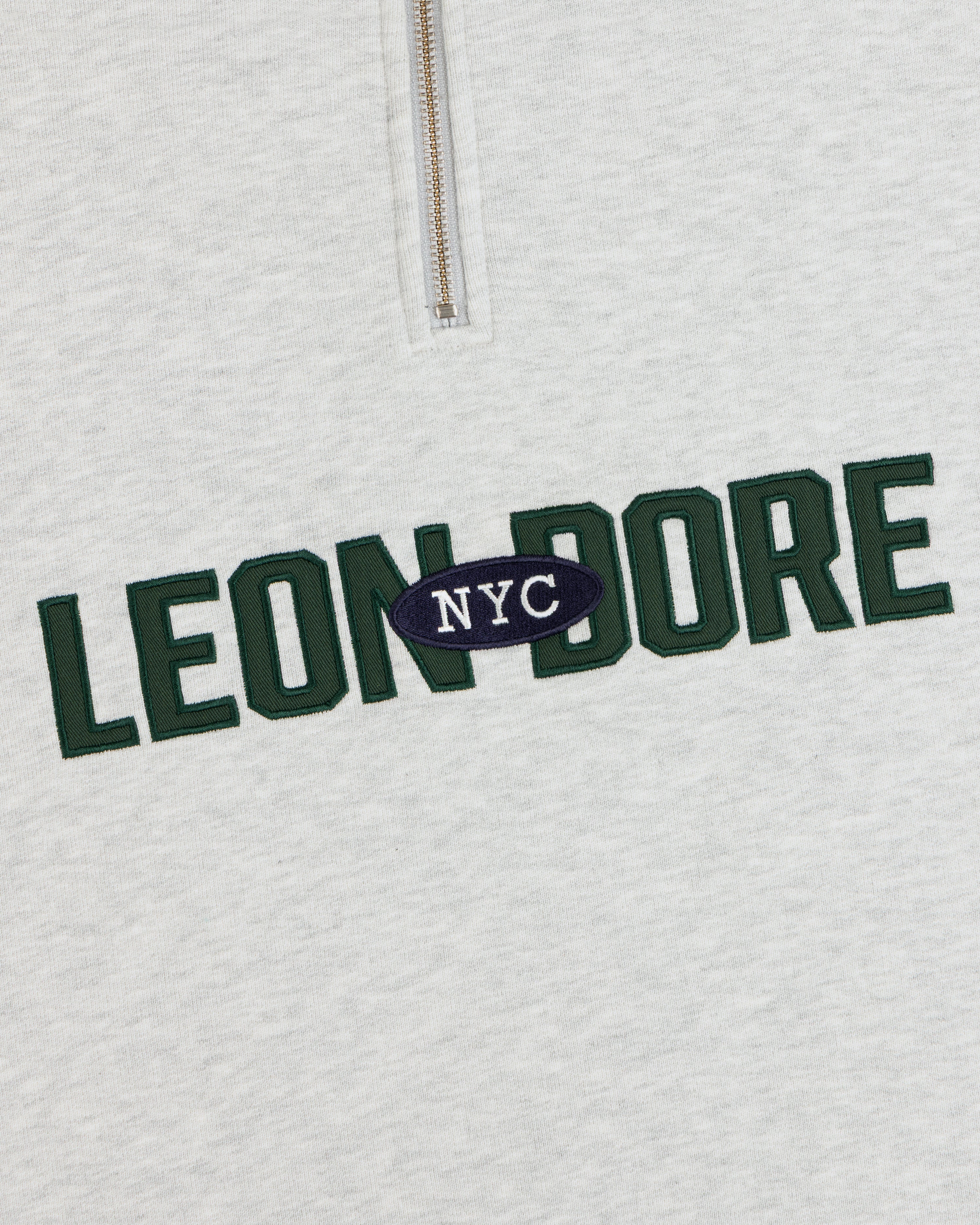 Close-up of Aimé Souvenir Quarter Zip’s light gray fabric, LEON DORE in green, NYC in white on a navy oval.