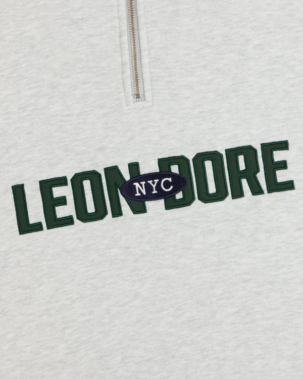 Close-up of Aimé Souvenir Quarter Zip’s light gray fabric, LEON DORE in green, NYC in white on a navy oval.